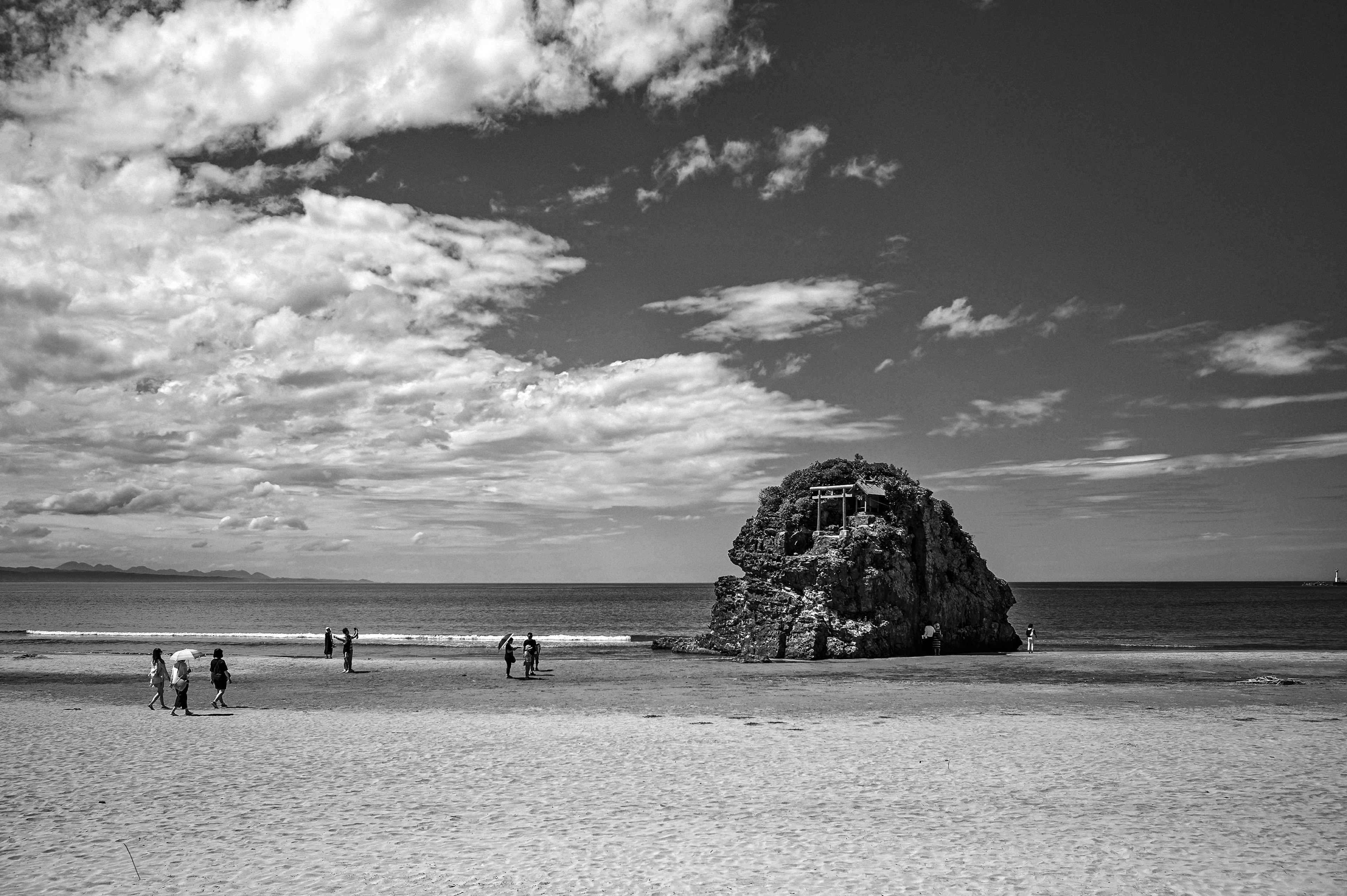 Large rock on a black and white beach with silhouettes of people