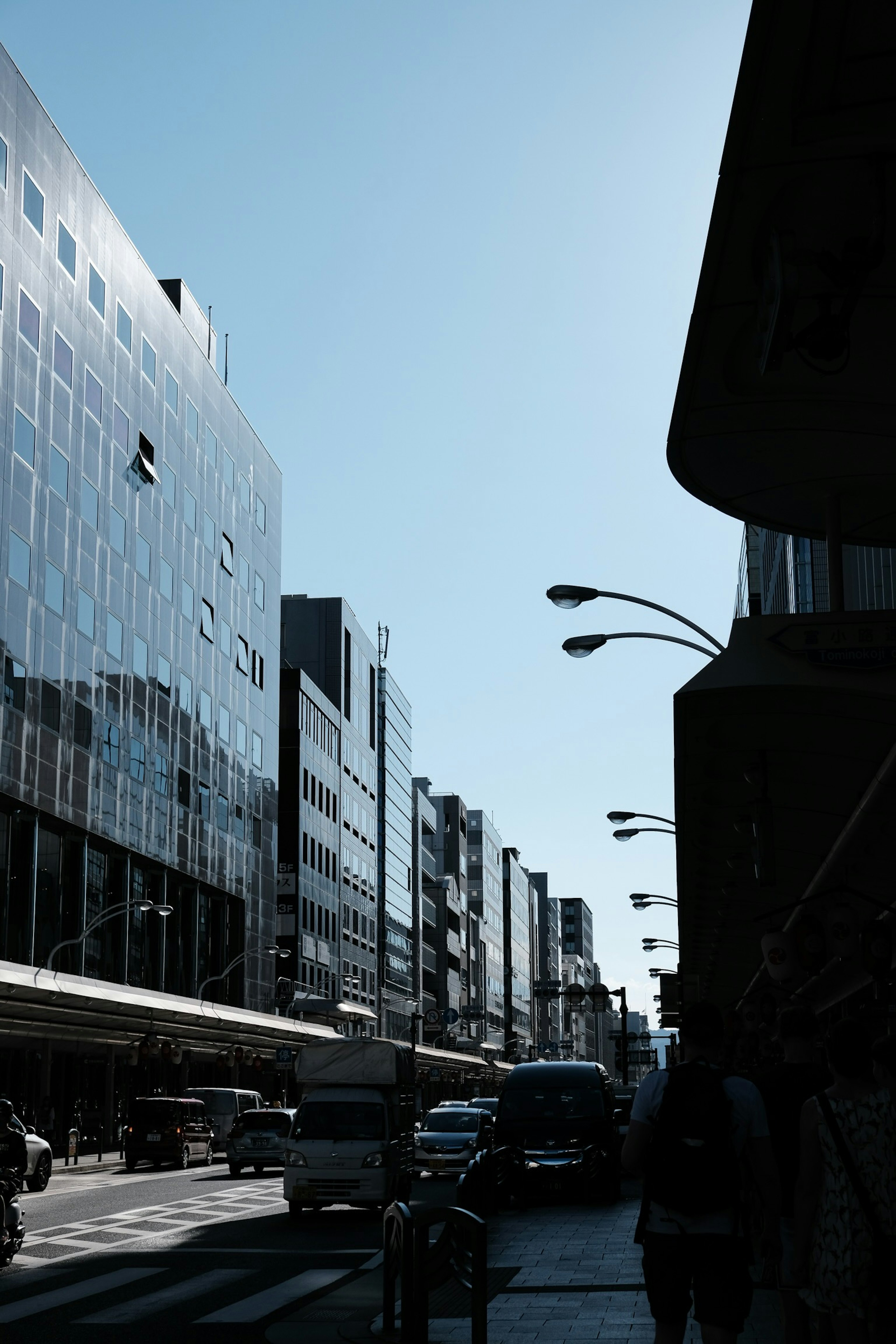 City street under a blue sky with modern glass buildings and visible traffic and pedestrians