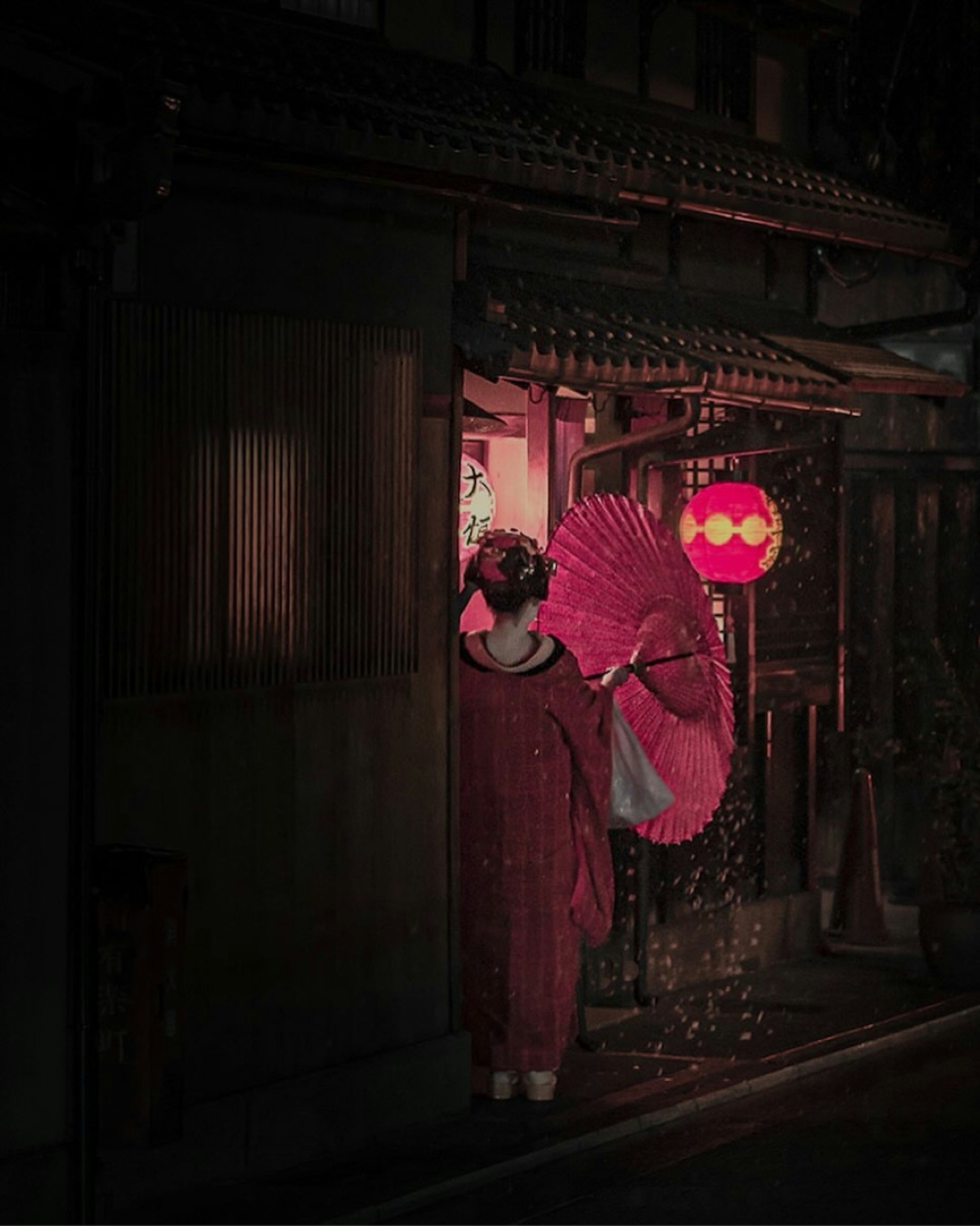 A woman in a red kimono holding an umbrella stands at a brightly lit entrance