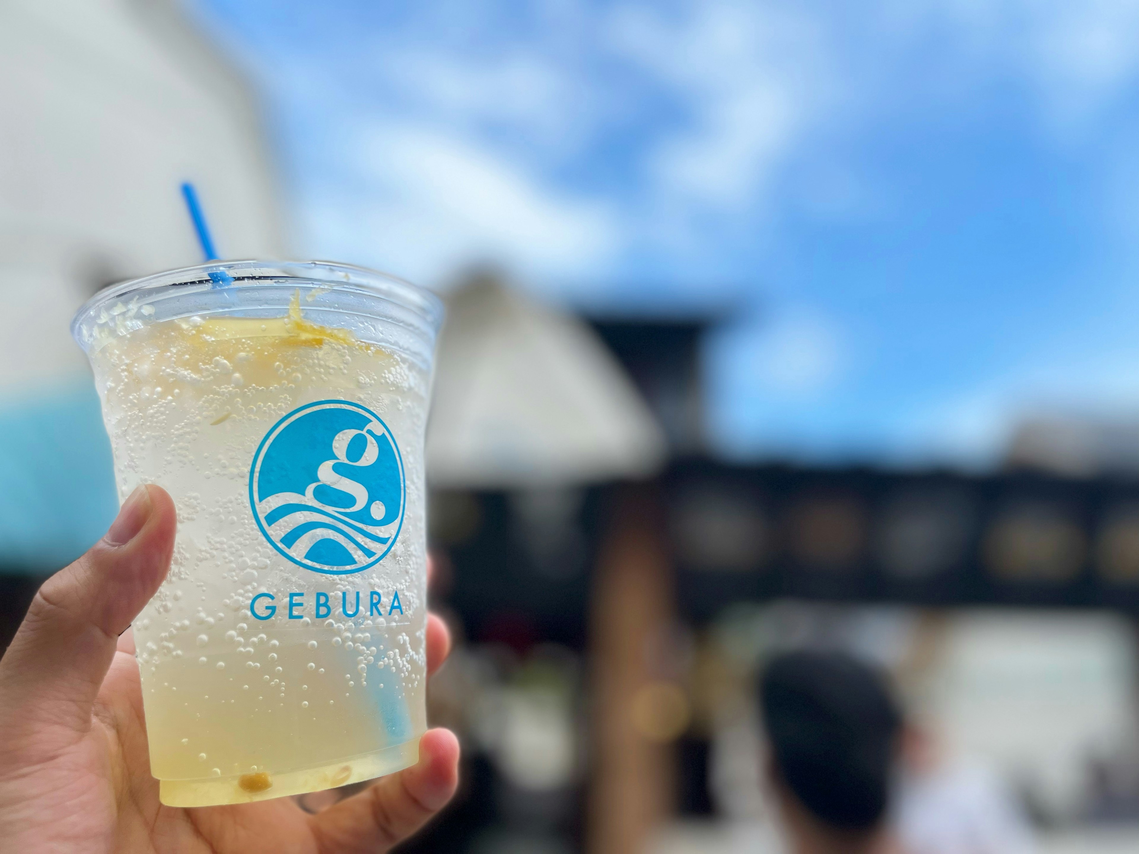 Una mano sosteniendo un vaso transparente de bebida con una pajilla azul contra un fondo de cielo azul y personas