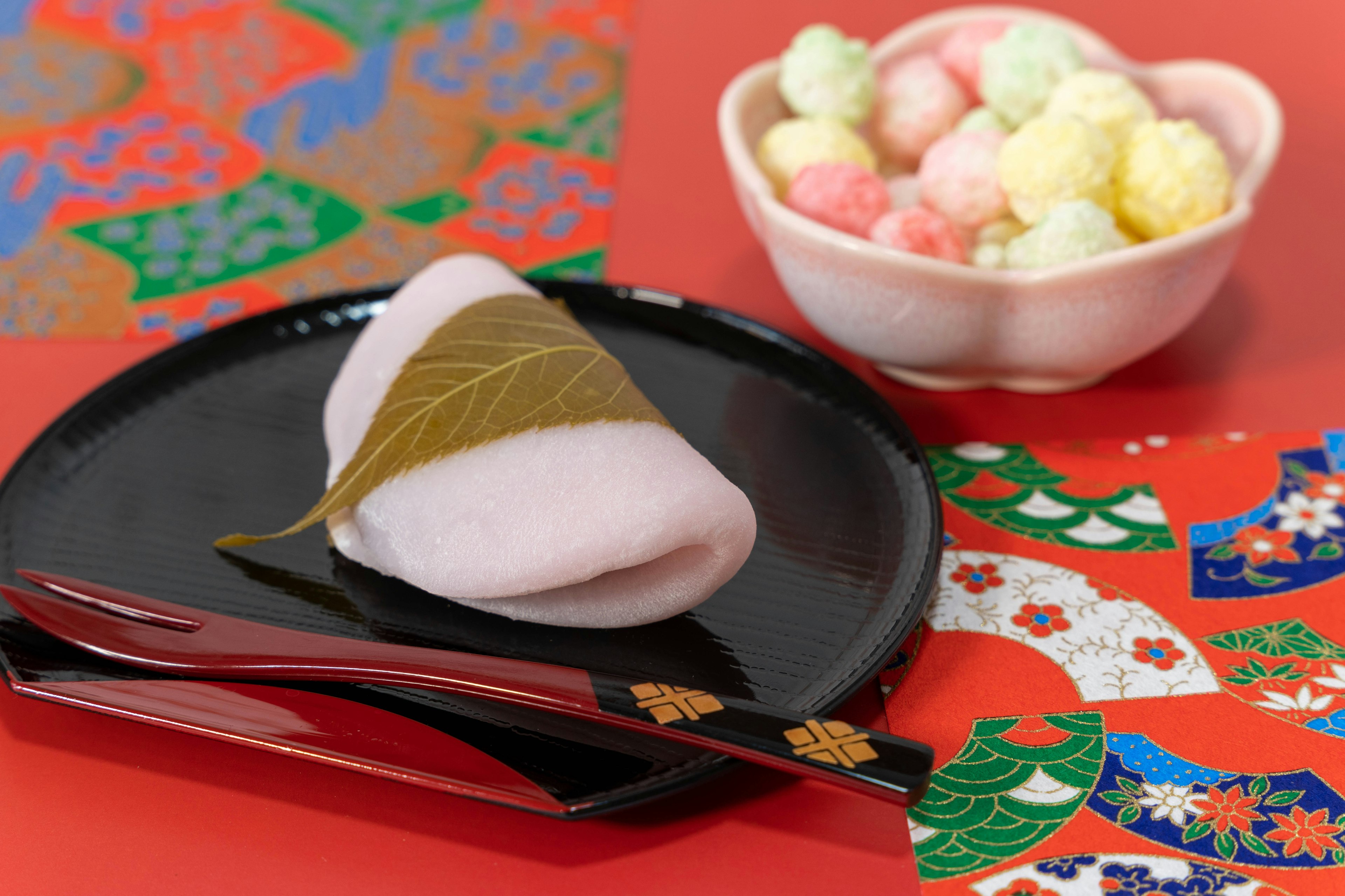 Sakura mochi placed on a black plate topped with a leaf, colorful Japanese patterned background