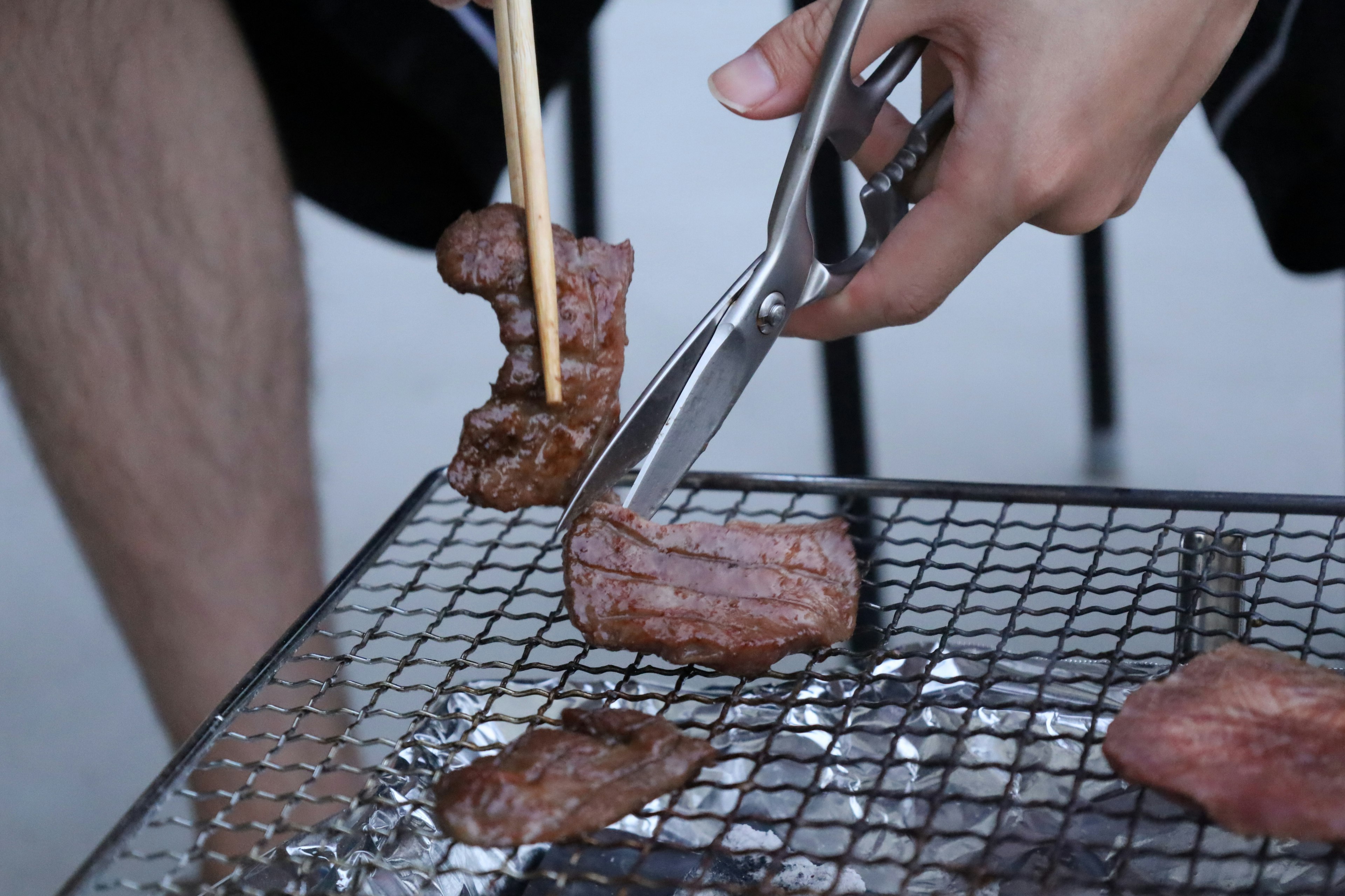 Una mano che tiene delle forbici e delle bacchette mentre taglia carne grigliata su una griglia