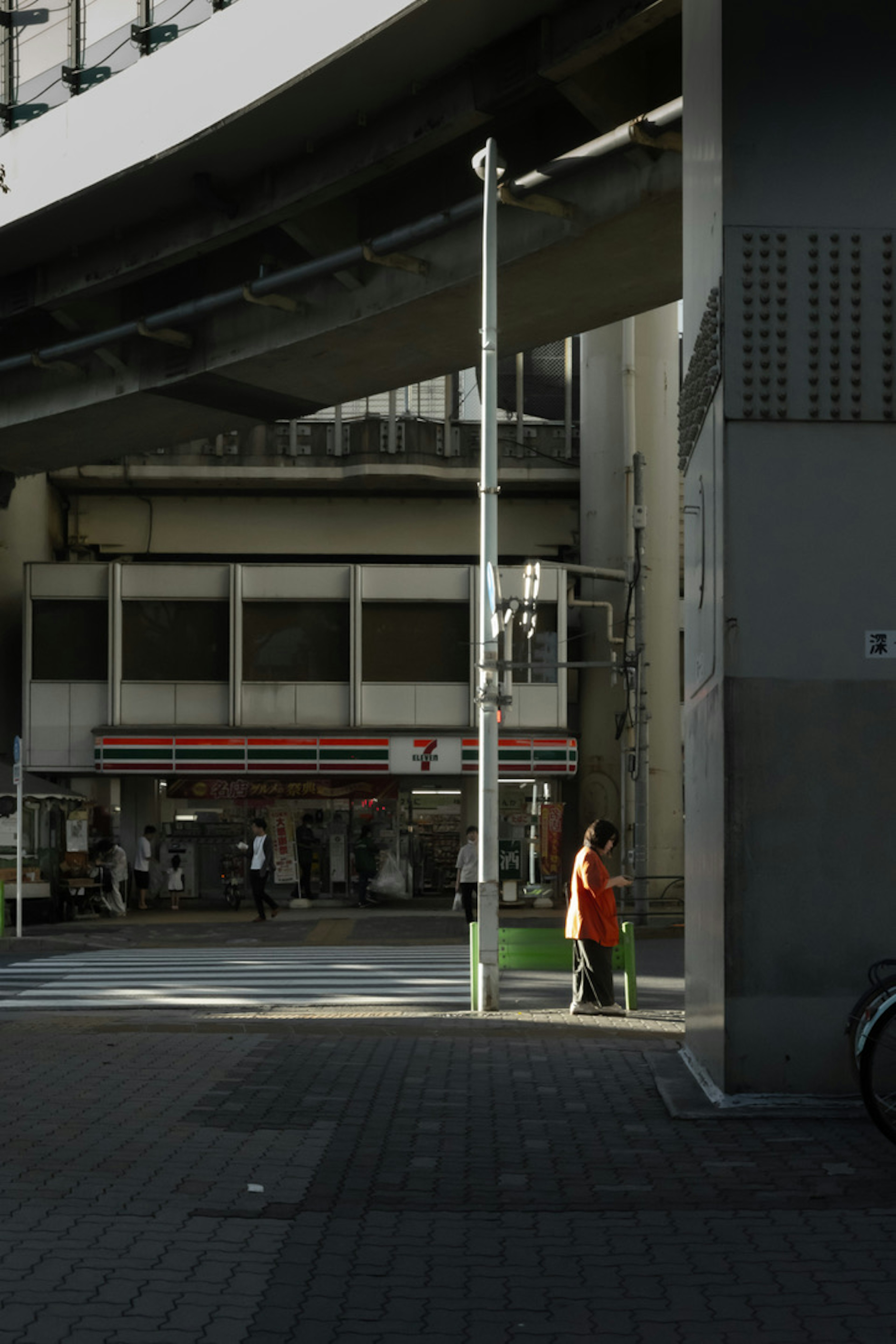 A woman in an orange outfit standing at an urban intersection with an elevated road