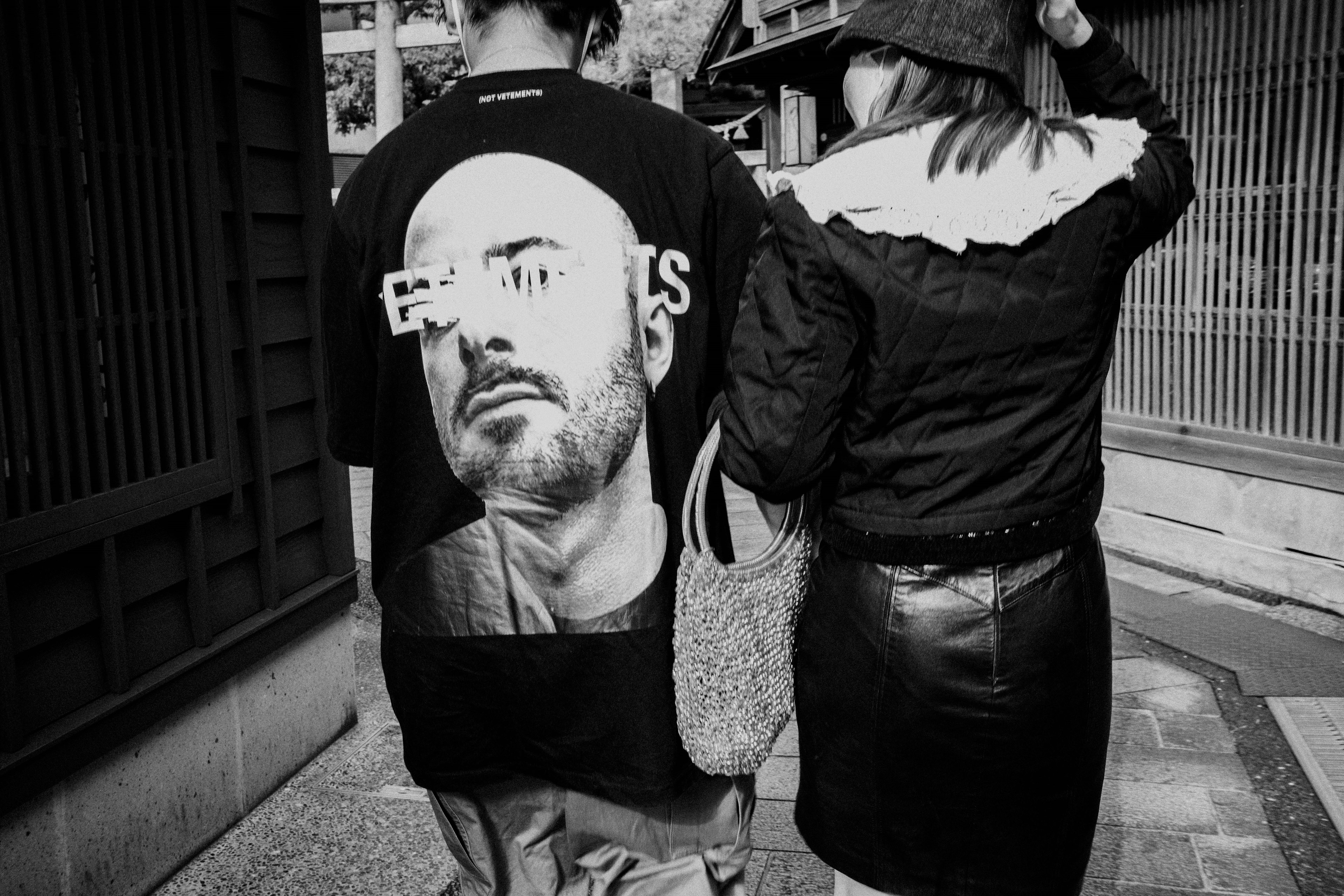 A monochrome image of two people walking with a large face print on the man's shirt