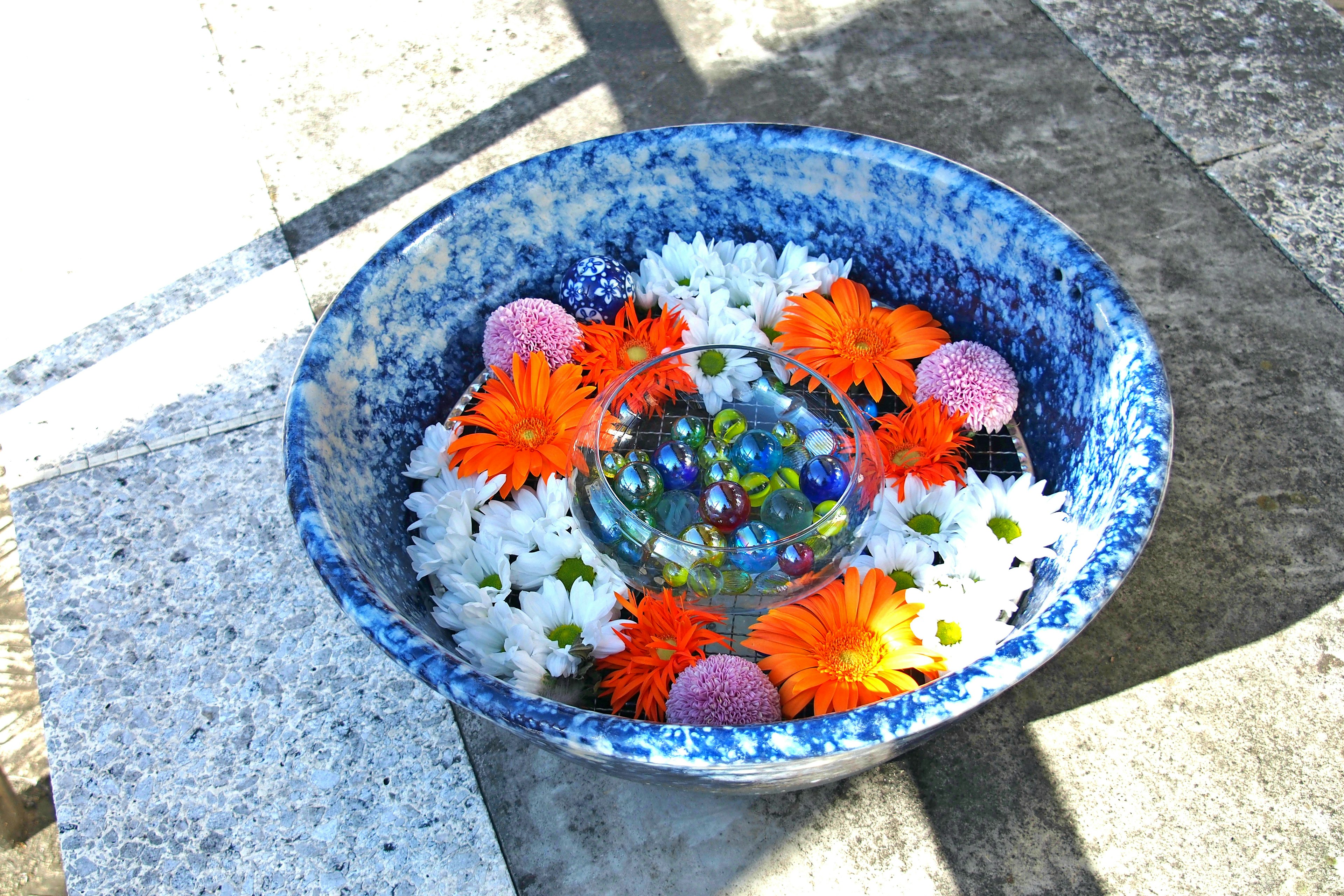 Un tazón azul lleno de flores blancas y naranjas adornado con canicas coloridas