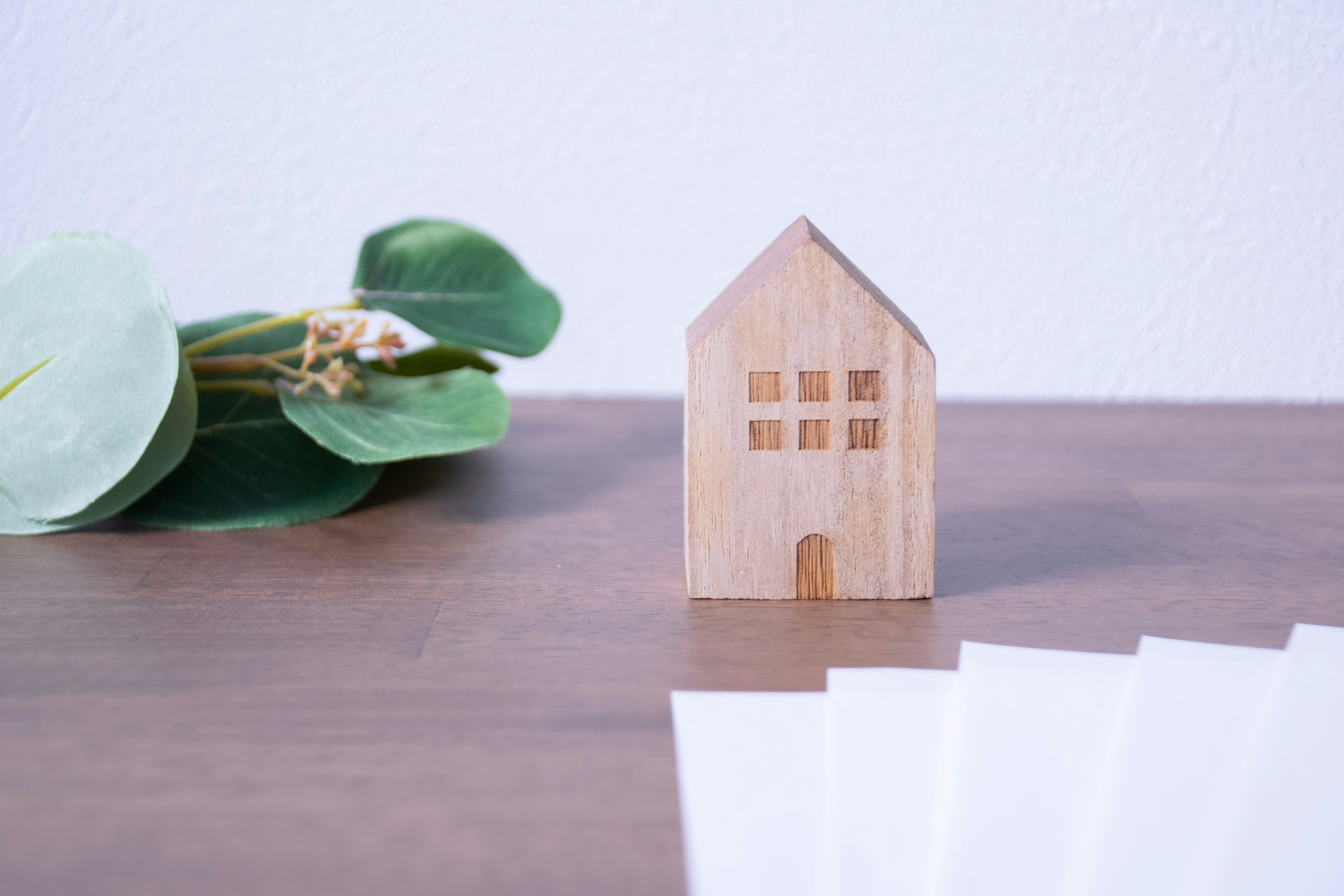 Un modello di casa in legno posato su un tavolo con fogliame verde e una pila di fogli bianchi accanto