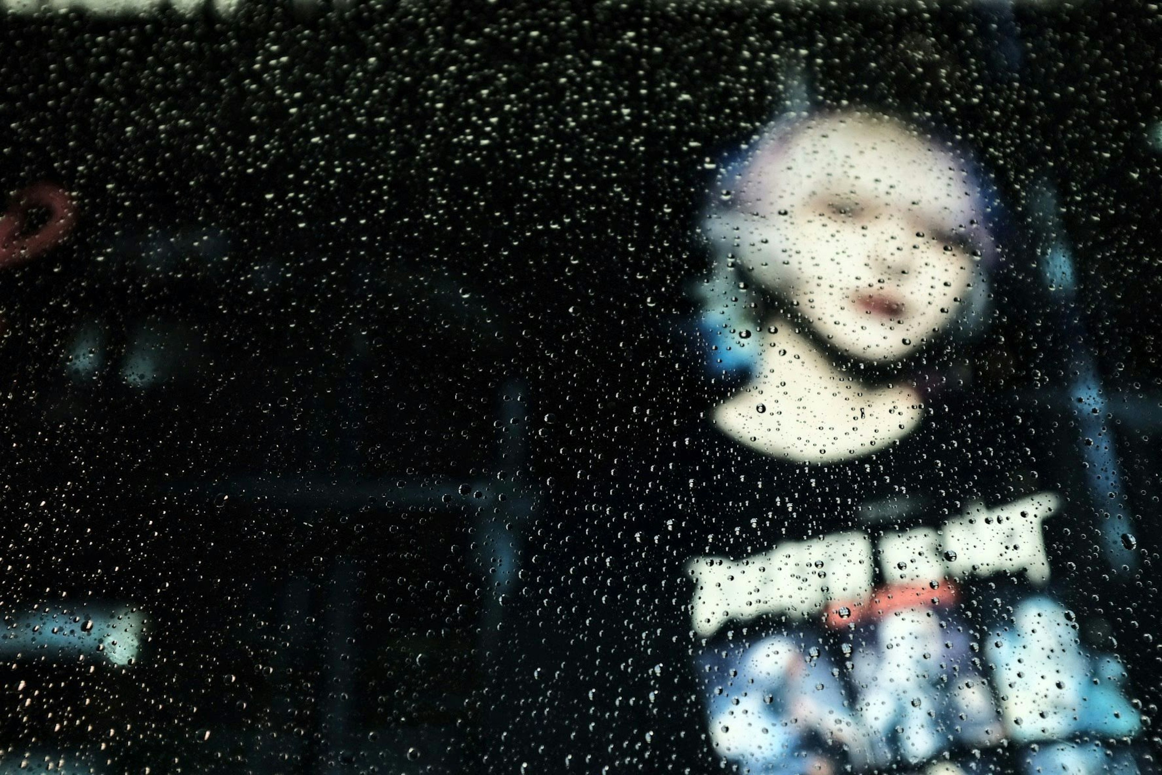 A blurred image of a woman behind a rain-speckled window featuring colorful hair and a black T-shirt