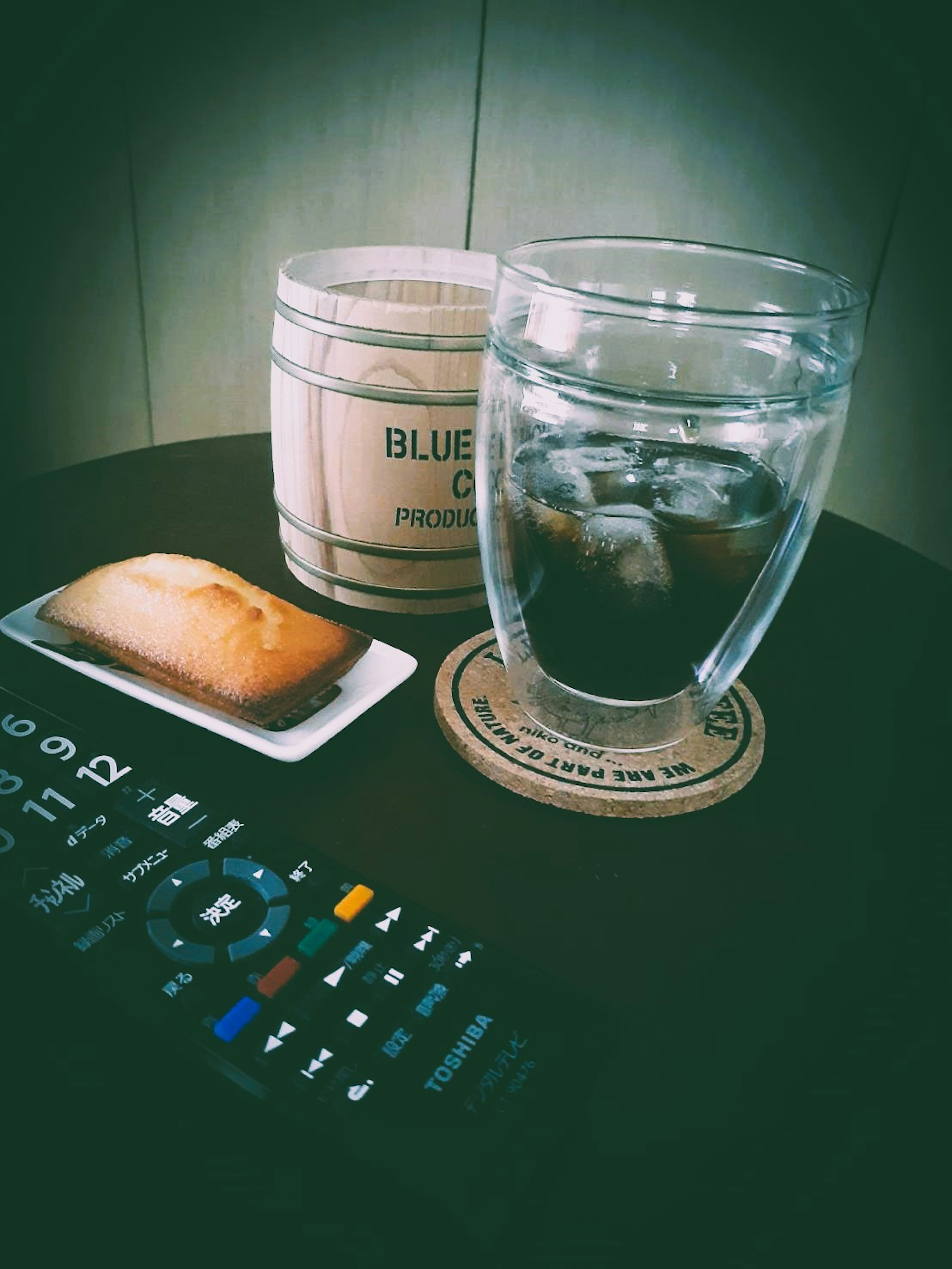 A glass with ice and a pastry on a table with a remote control and a coaster