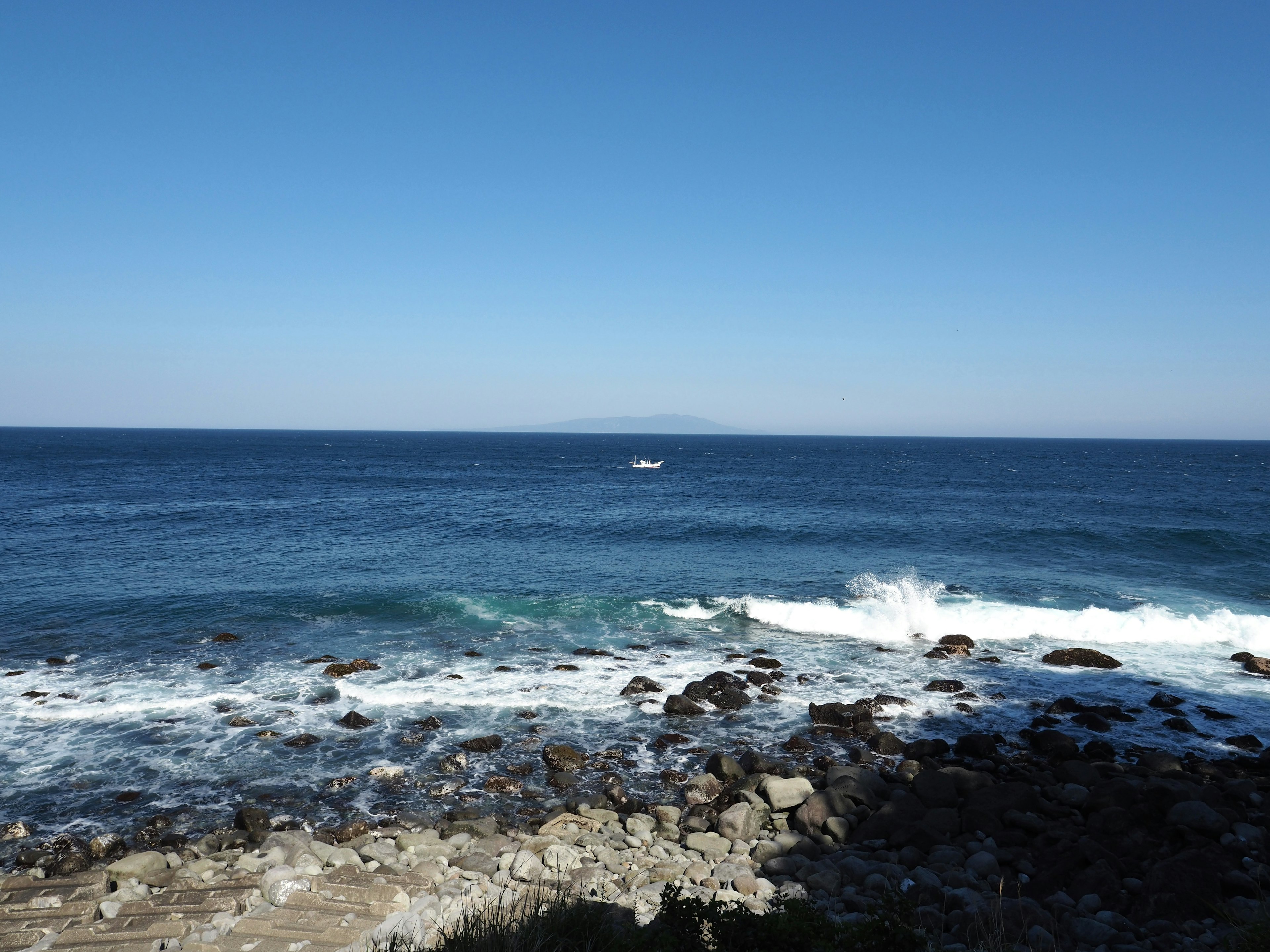 Landschaft mit blauem Ozean und weißen Wellen sowie einem kleinen Boot an der felsigen Küste