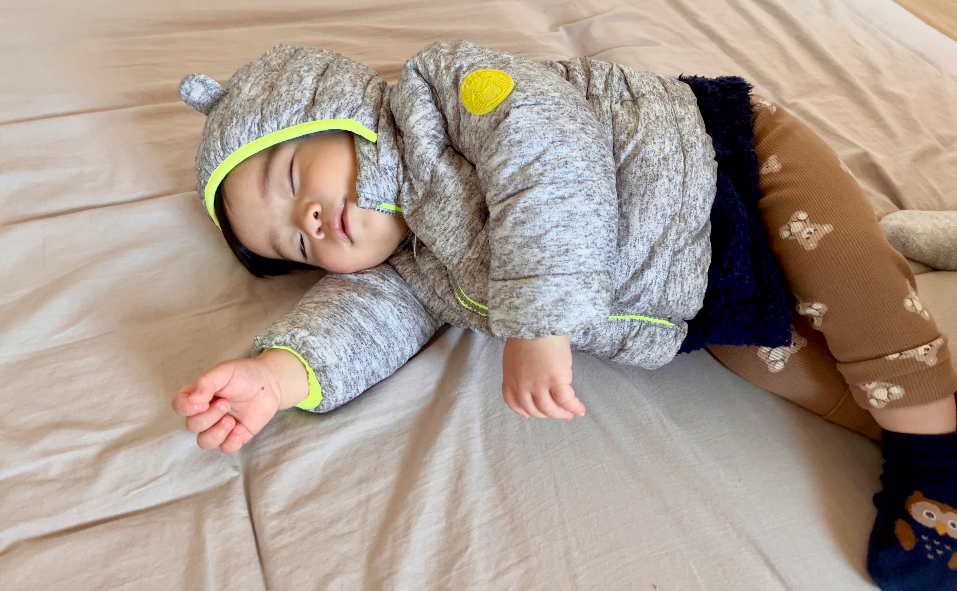 A baby wearing a gray hooded jacket sleeping on a bed