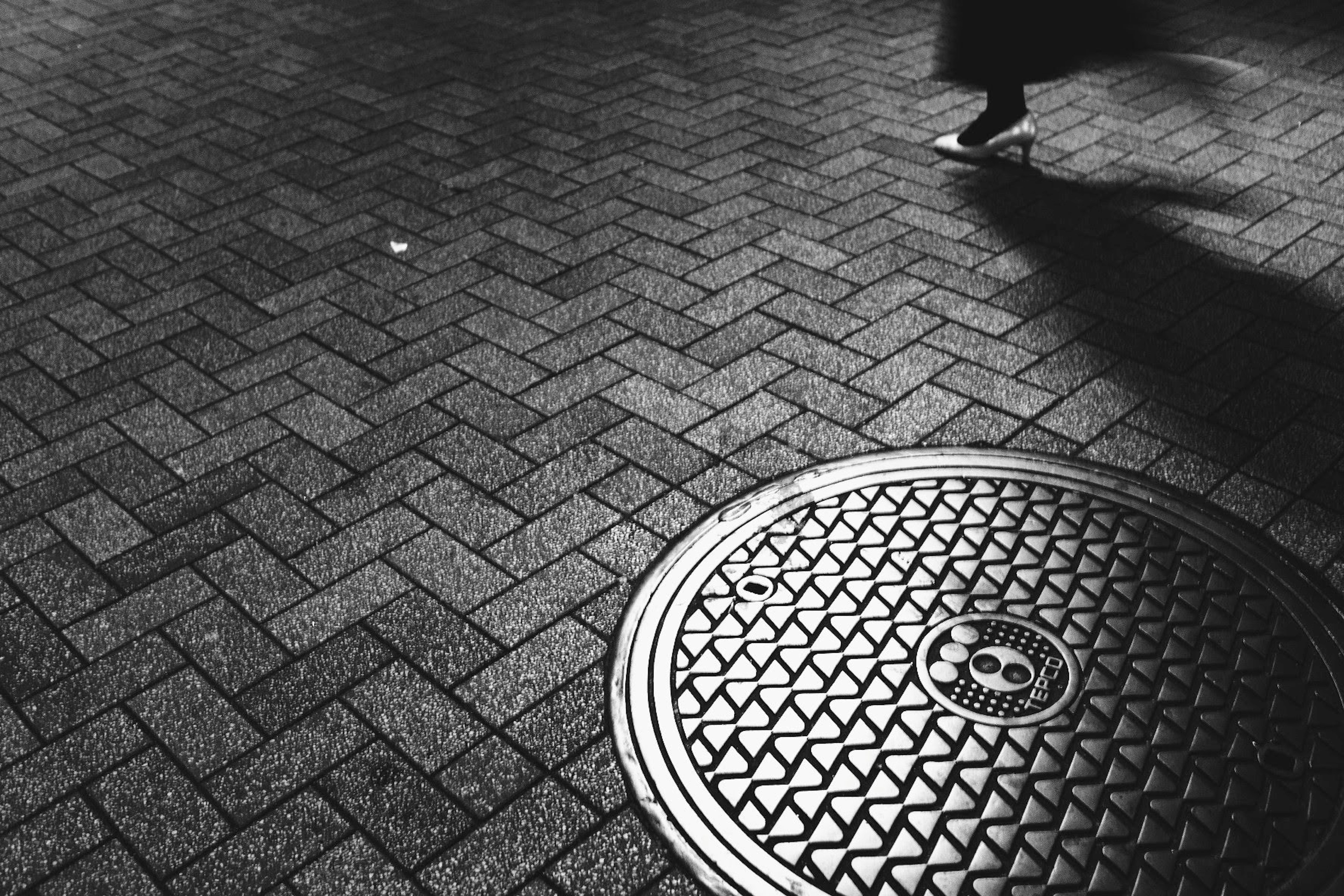 Escena urbana en blanco y negro con una tapa de alcantarilla y los pies de un peatón