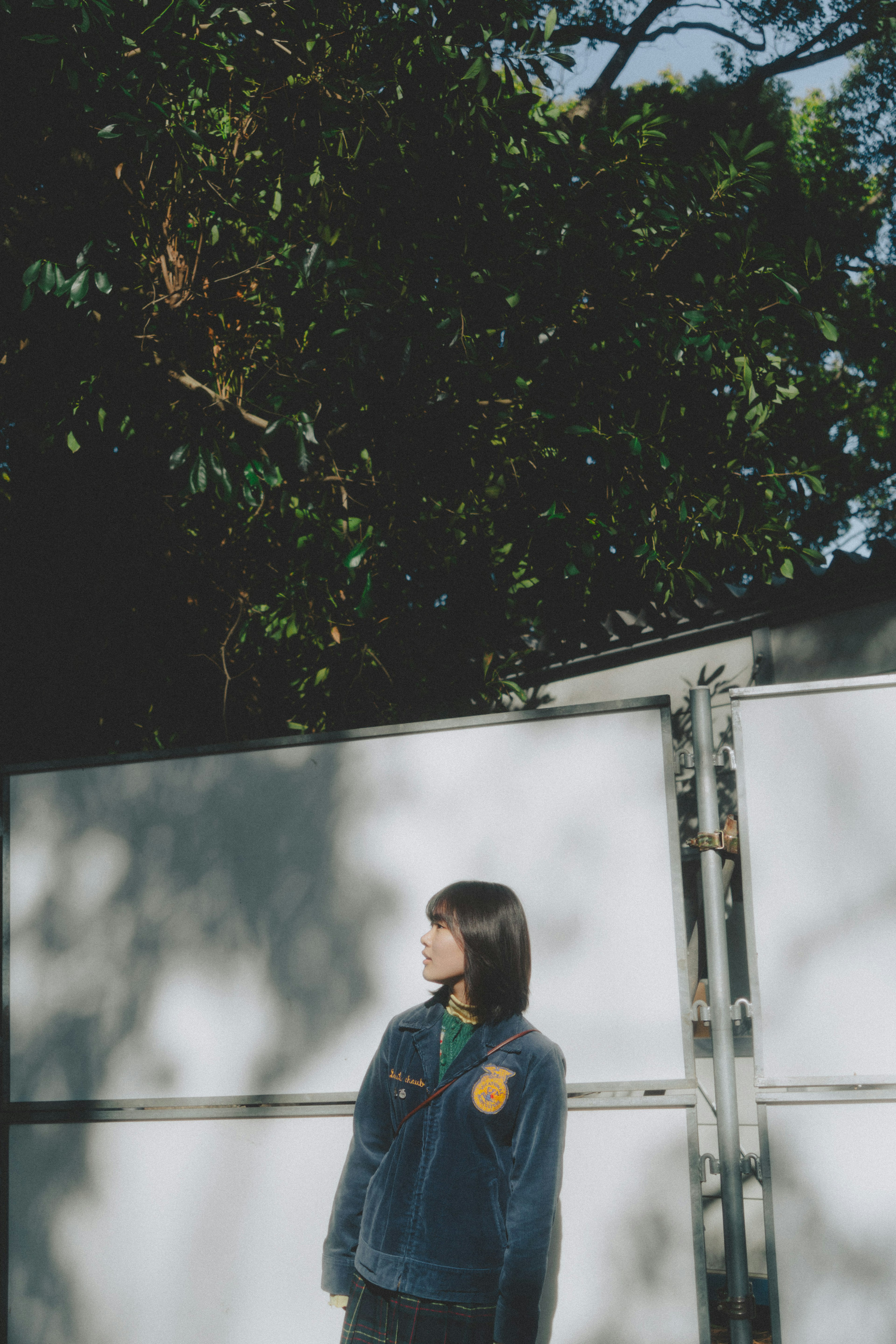 A young woman wearing a denim jacket standing in front of a white fence