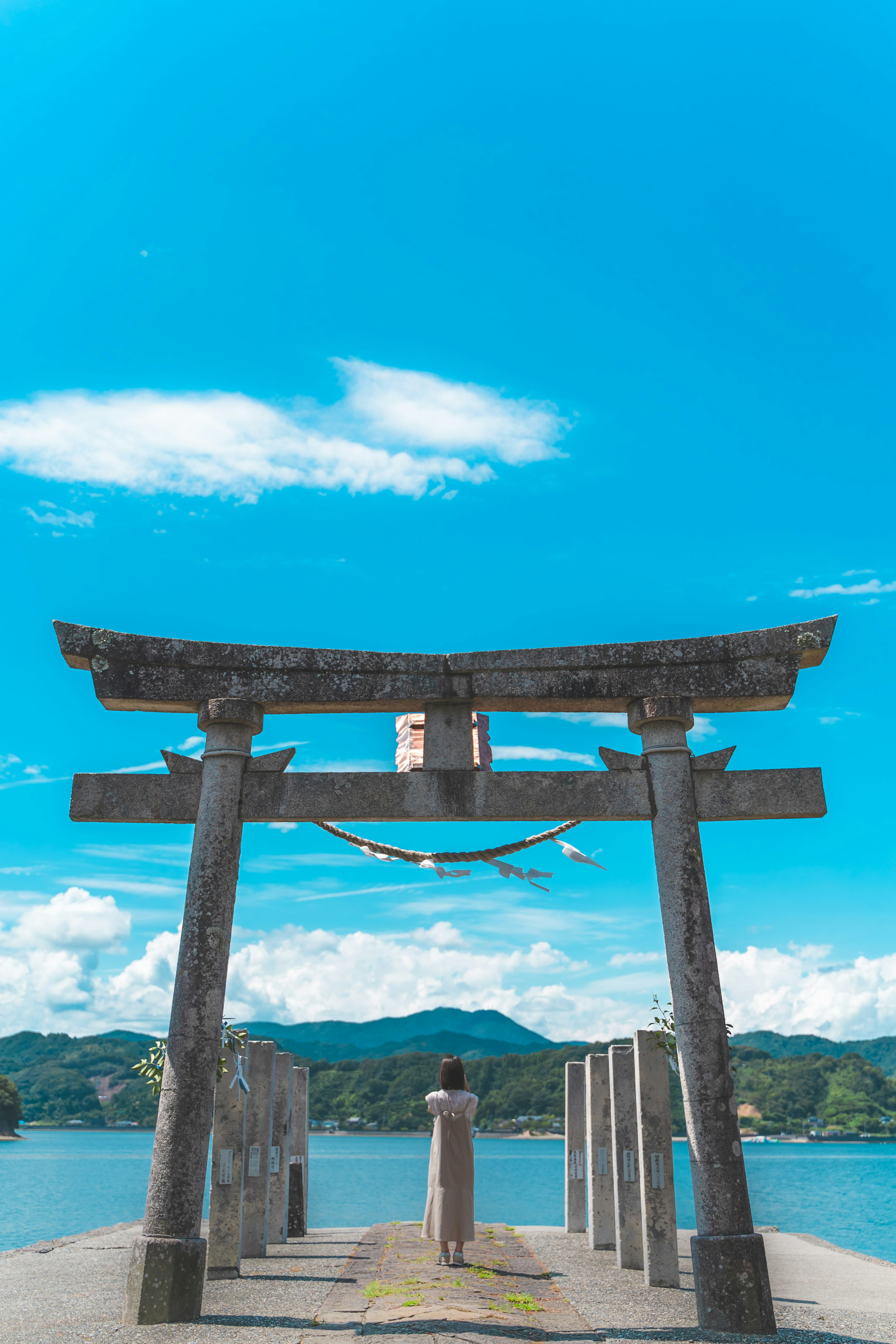 青空の下にある鳥居の写真で、静かな海と山々が背景に見える