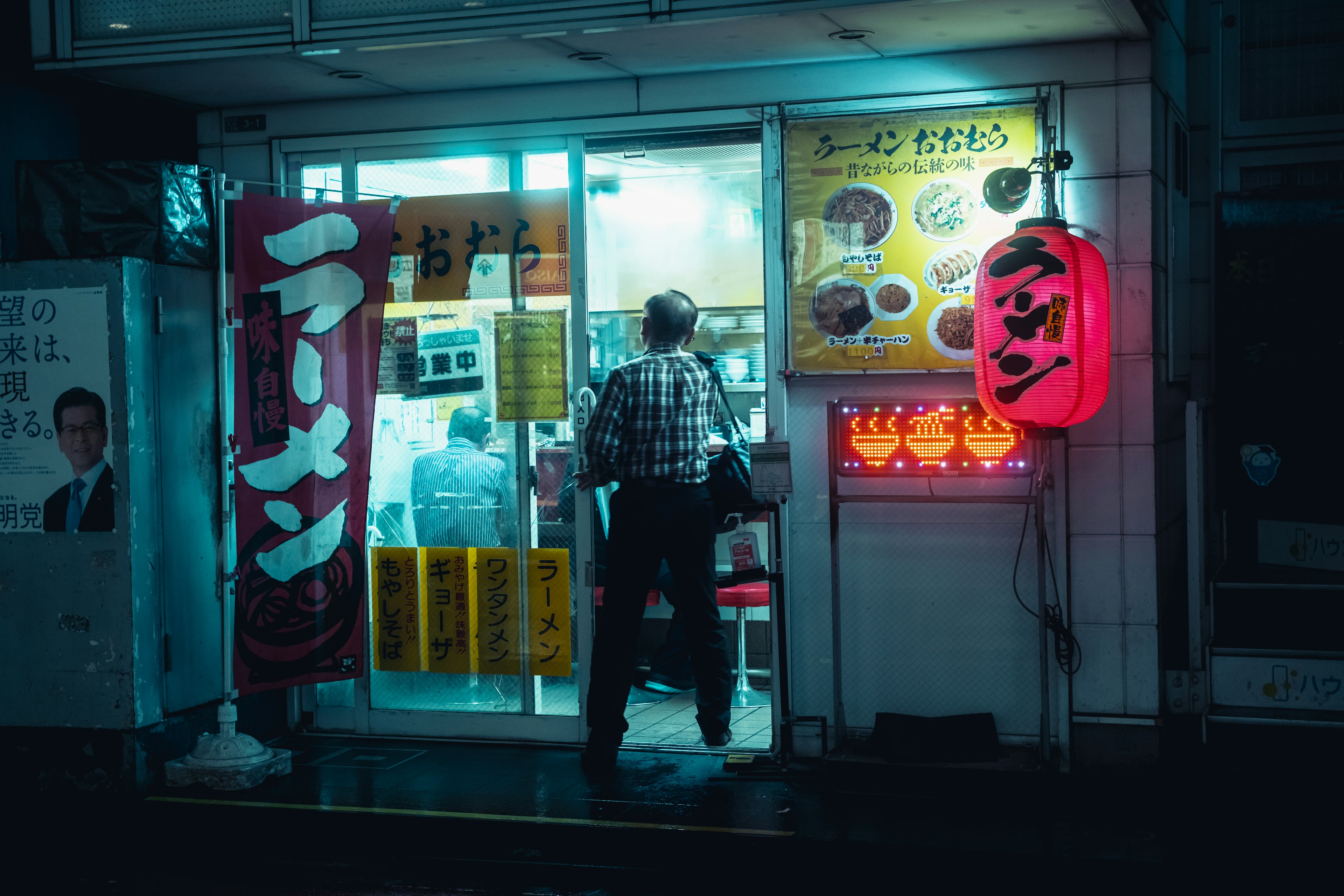 Exterior de una tienda de ramen iluminada por luz azul con un cliente sosteniendo una linterna roja