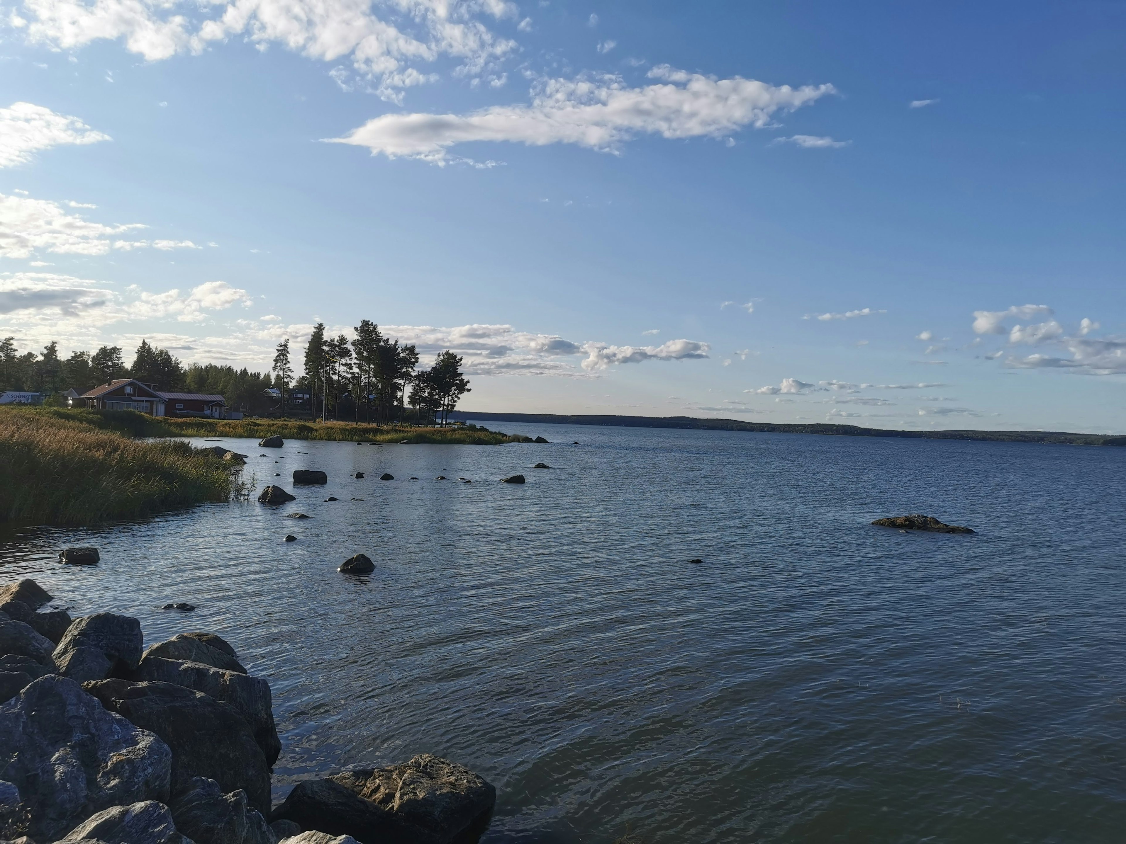 Serene lake view with blue sky and white clouds featuring rocks and grass along the shore