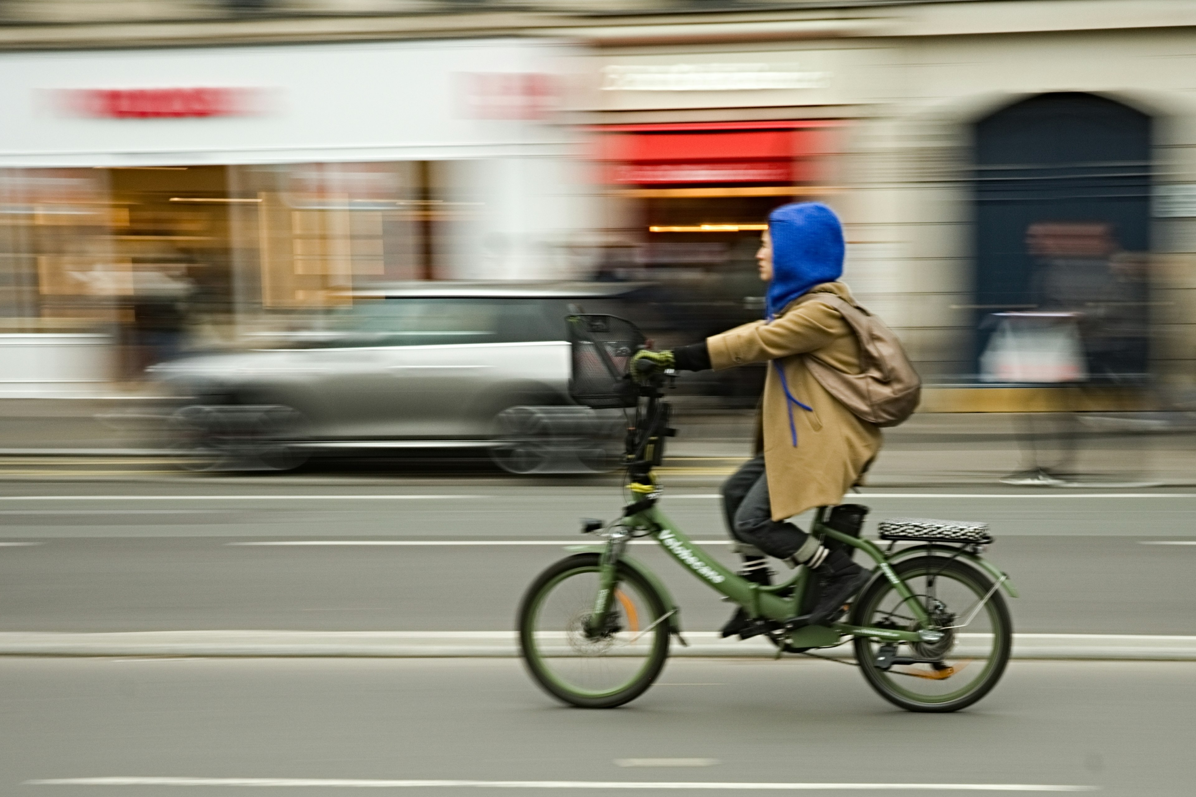 A person wearing a blue hood rides a bicycle in a dynamic urban setting