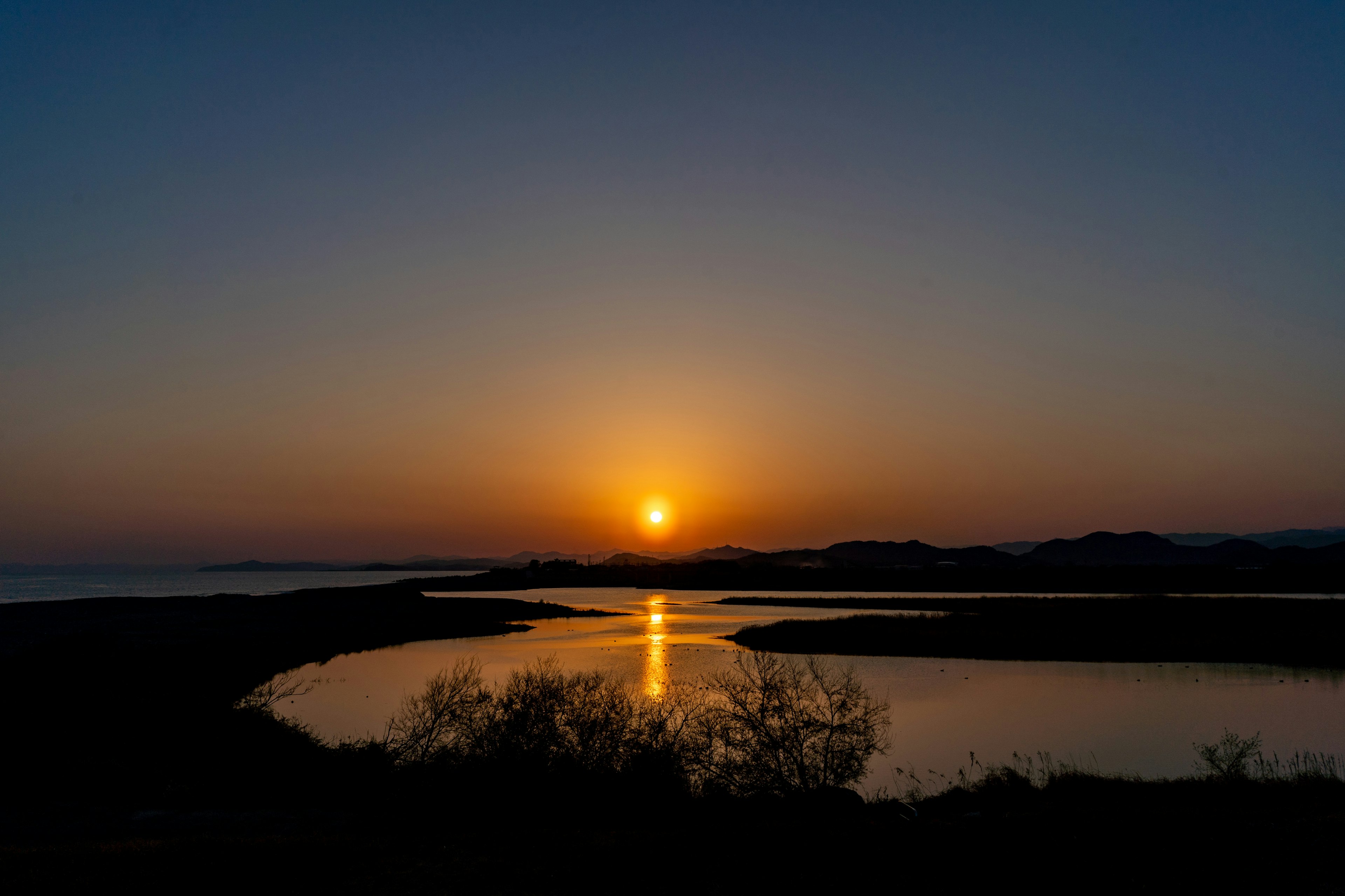 A serene sunset over a calm water body with a vibrant orange sun and a blue sky