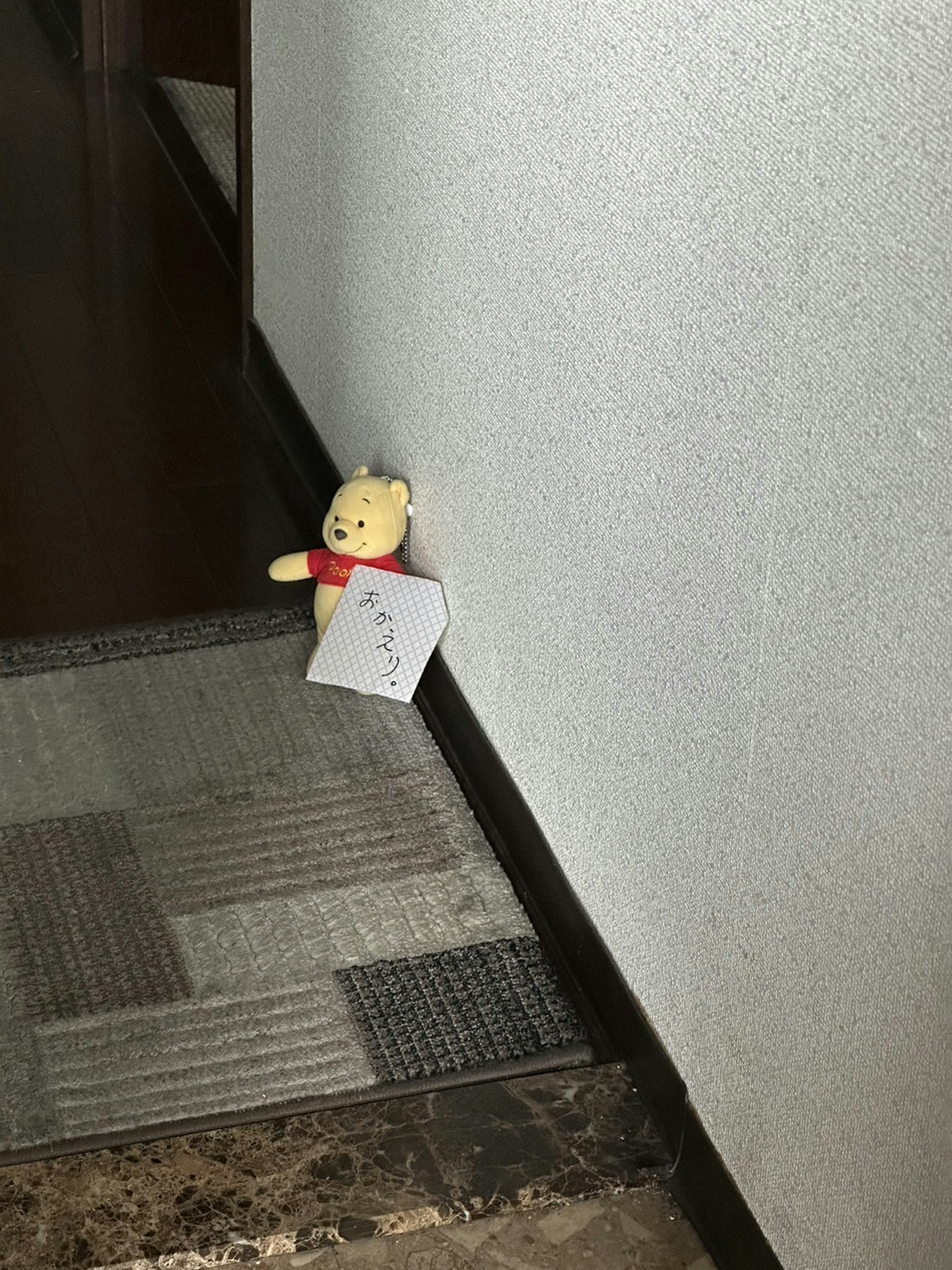 A small teddy bear placed in the corner of a hallway holding a note