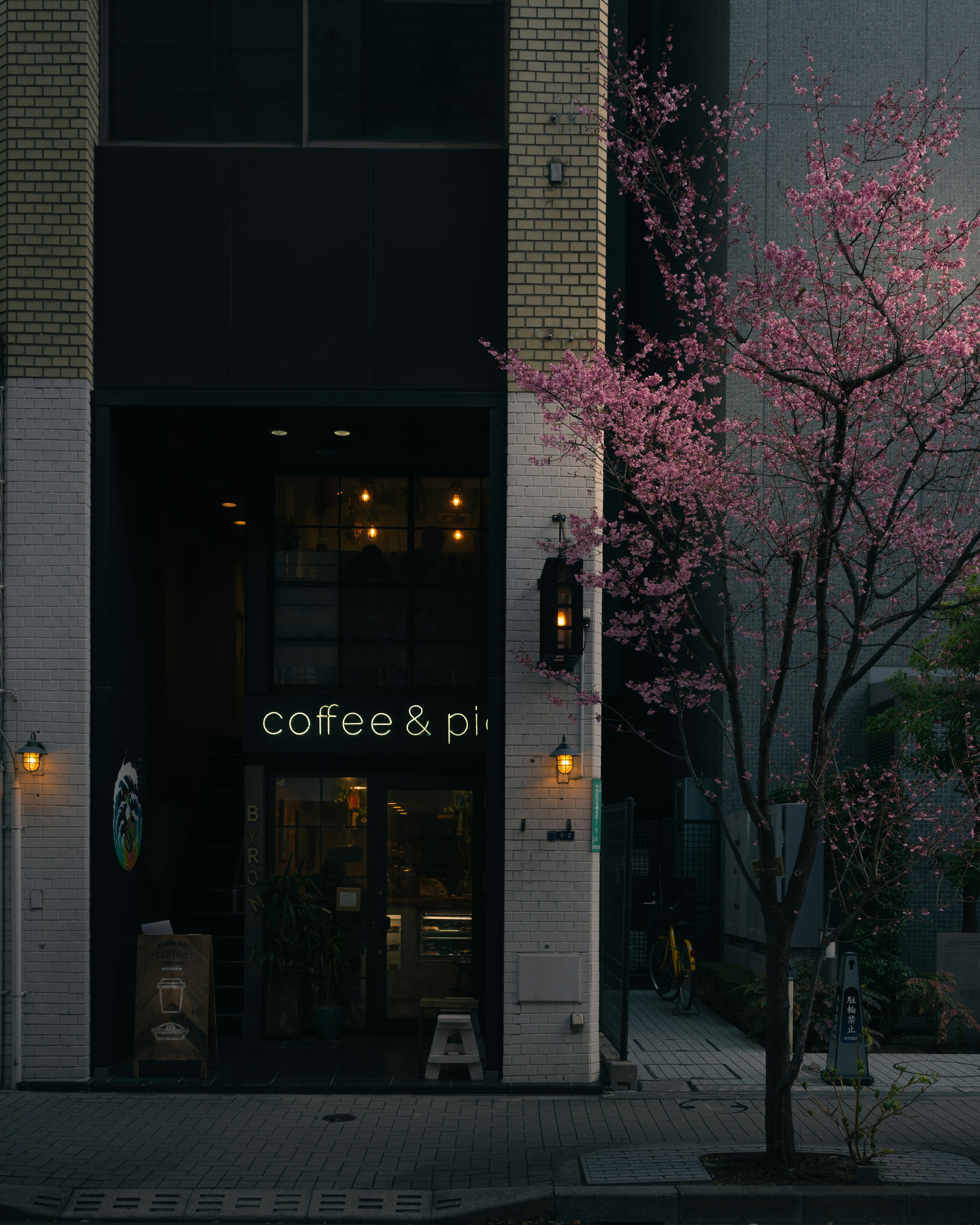 Fachada de una cafetería con un árbol en flor rosa y un letrero de café y pizza