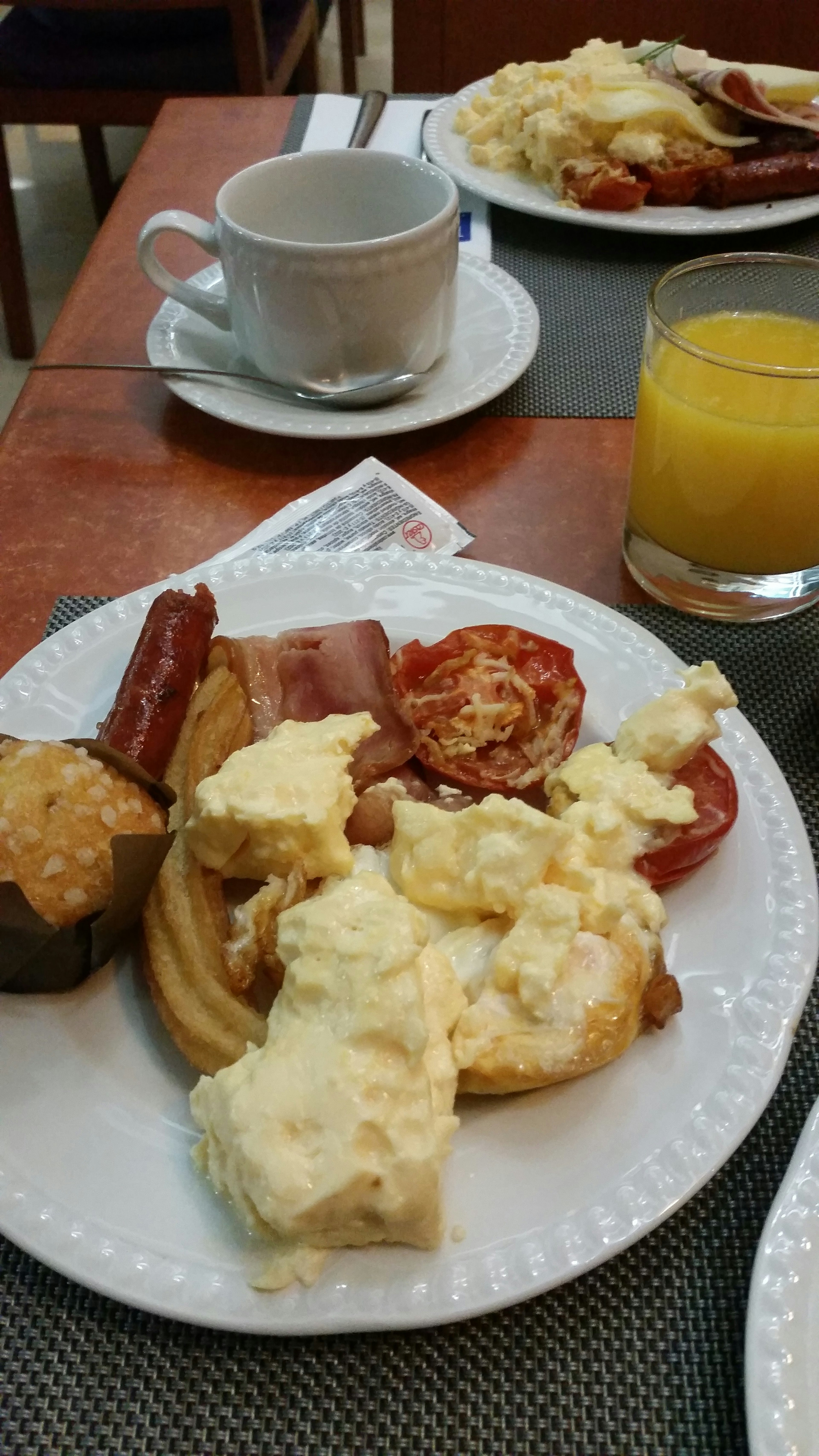 Plato de desayuno con huevos, salchichas, tocino, tomates, muffin y jugo de naranja