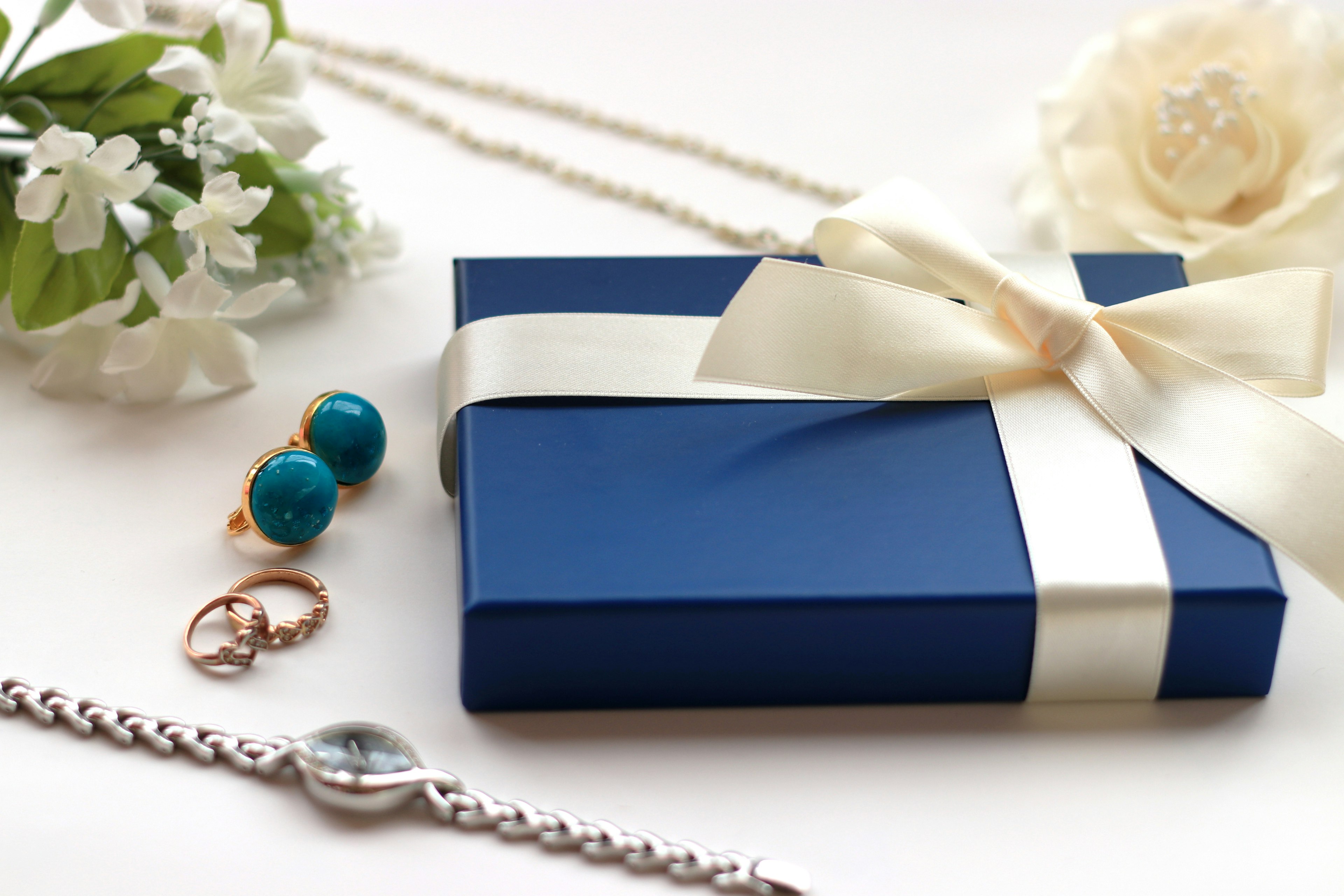 Blaue Geschenkbox mit weißem Band umgeben von Schmuck Silbernes Armband und Ringe Blaue Ohrringe und Halskette Weiße Blume