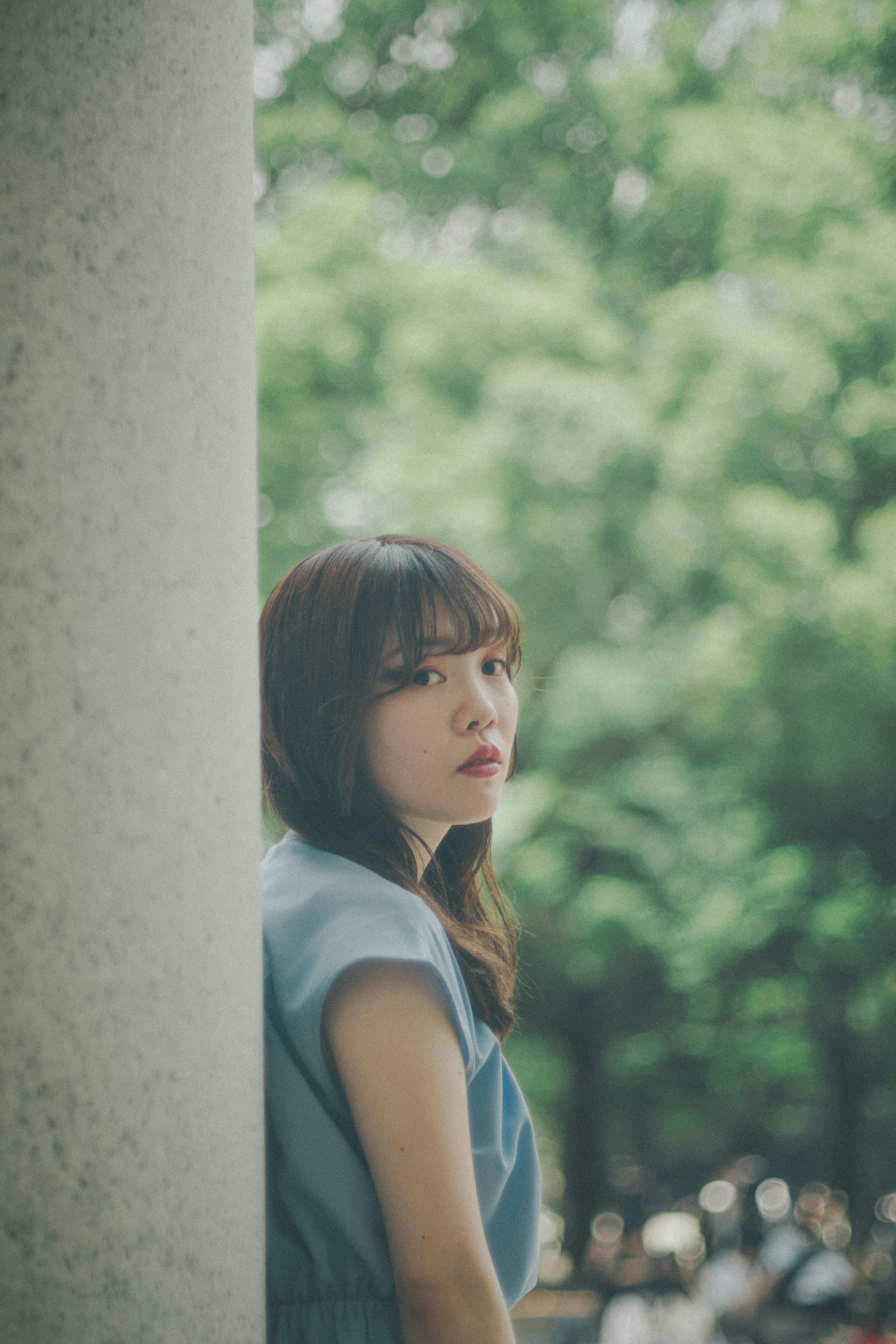 A woman in a blue dress leaning against a pillar gazing outdoors with a green background