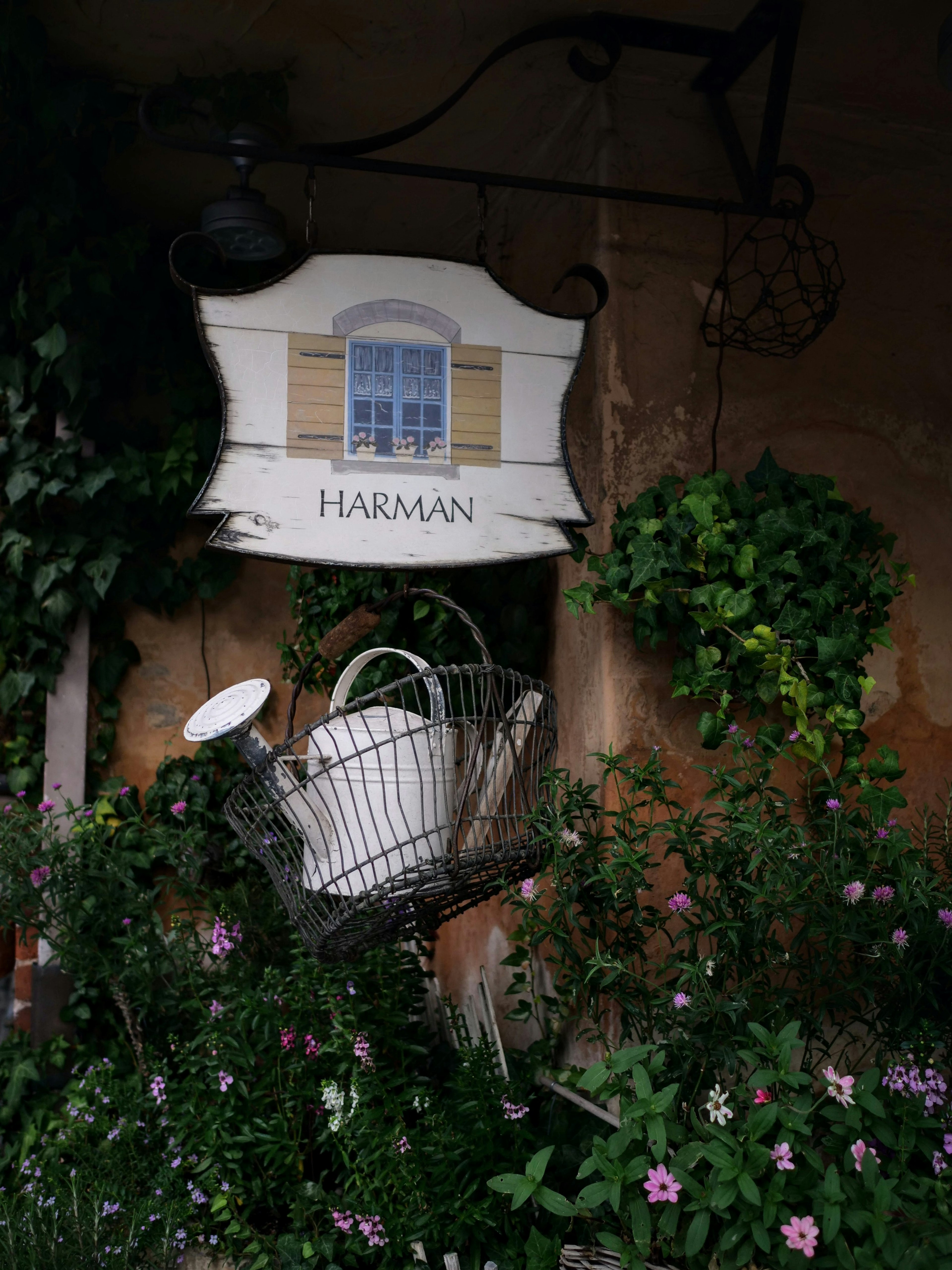 Eine weiße Gießkanne und ein Schild mit der Aufschrift HARMAN in einem blühenden Garten