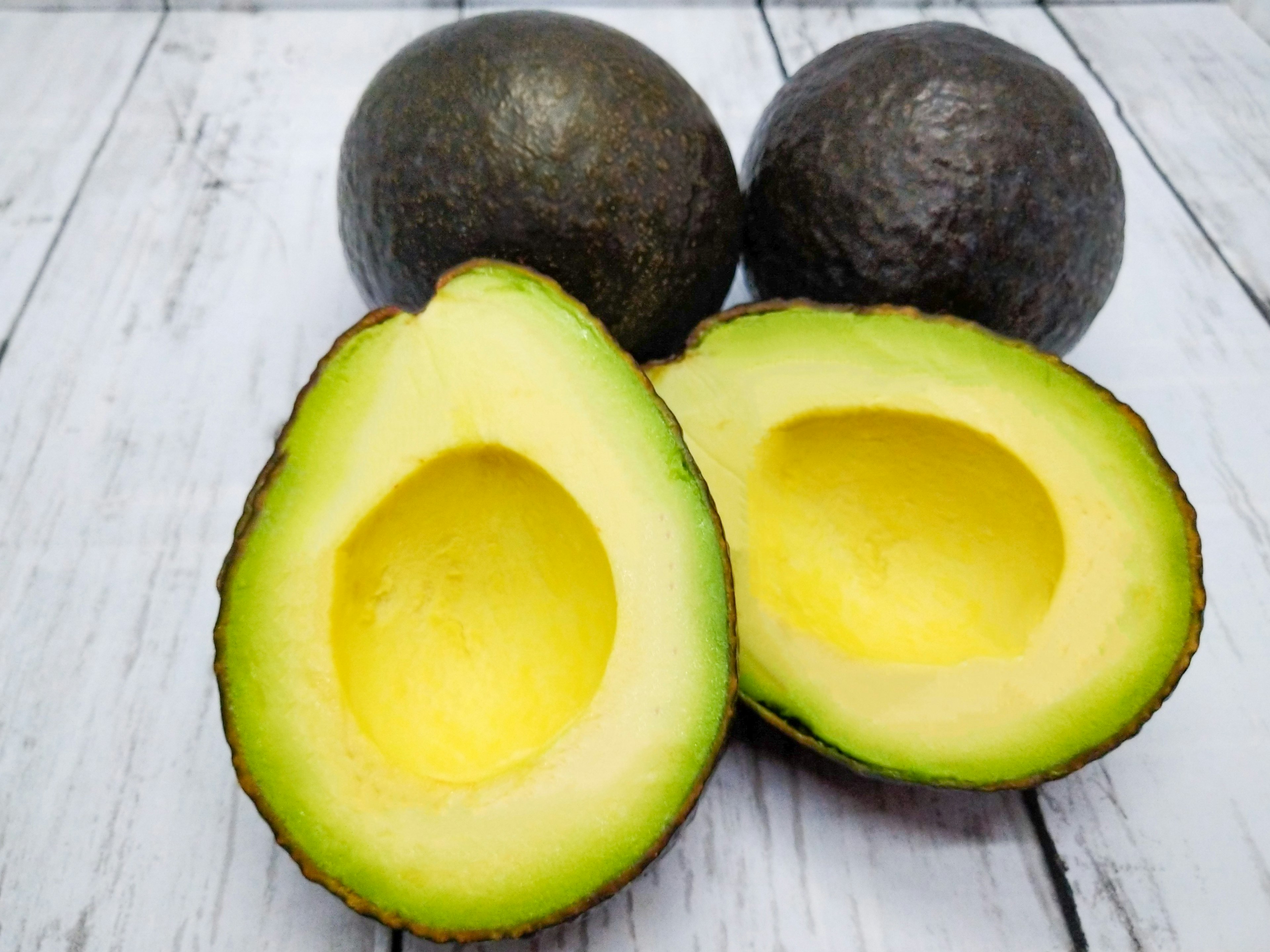 Halved avocado with bright yellow flesh next to whole avocados on a wooden surface