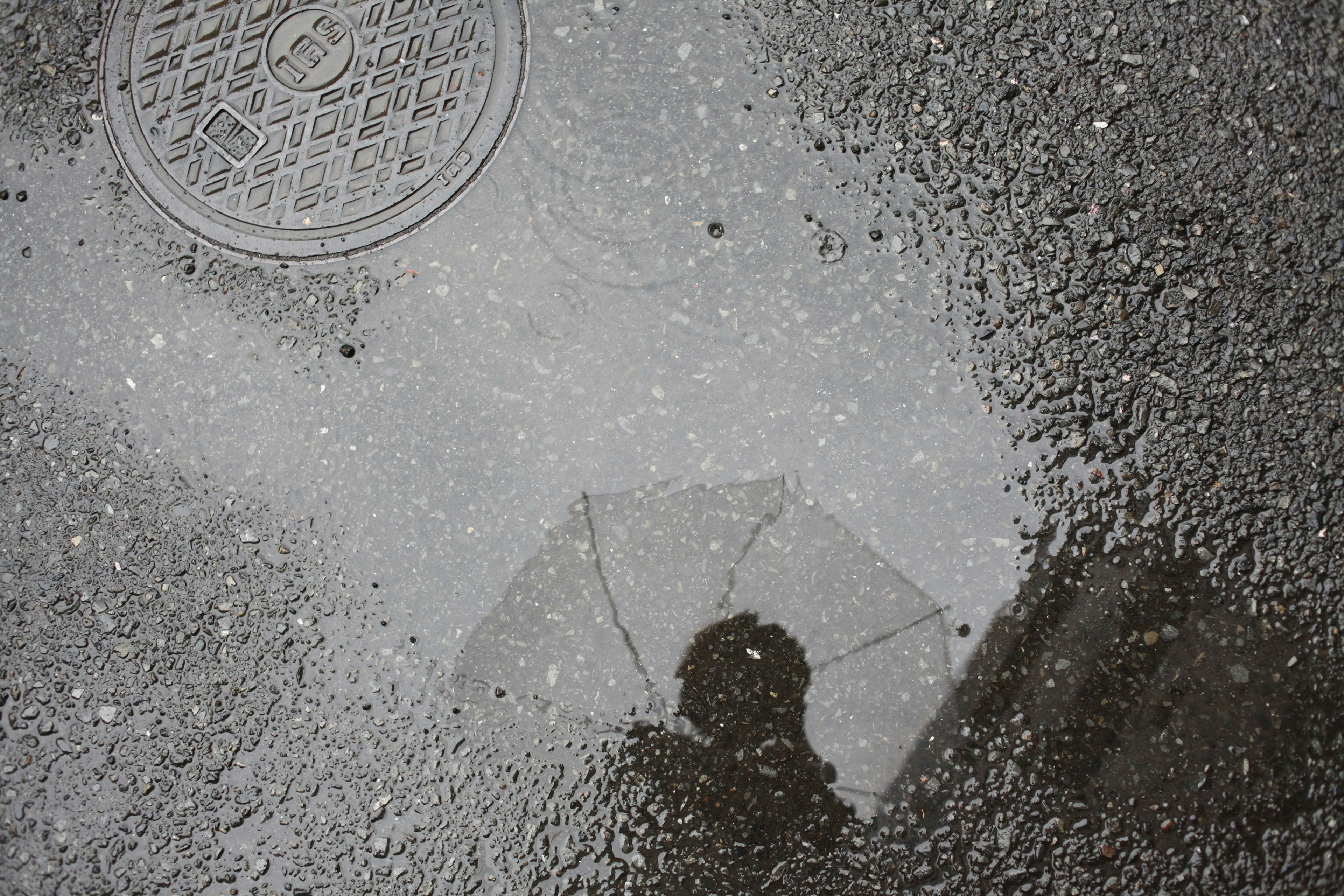 Reflection of a person with an umbrella in a puddle alongside a manhole cover