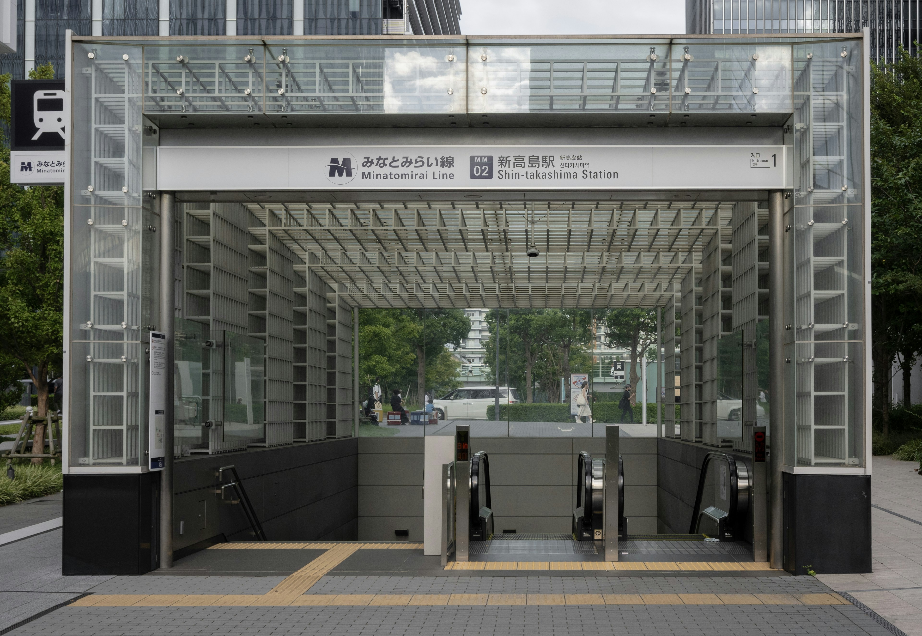 Entrée de métro au design transparent donnant sur une verdure urbaine divers bâtiments en arrière-plan