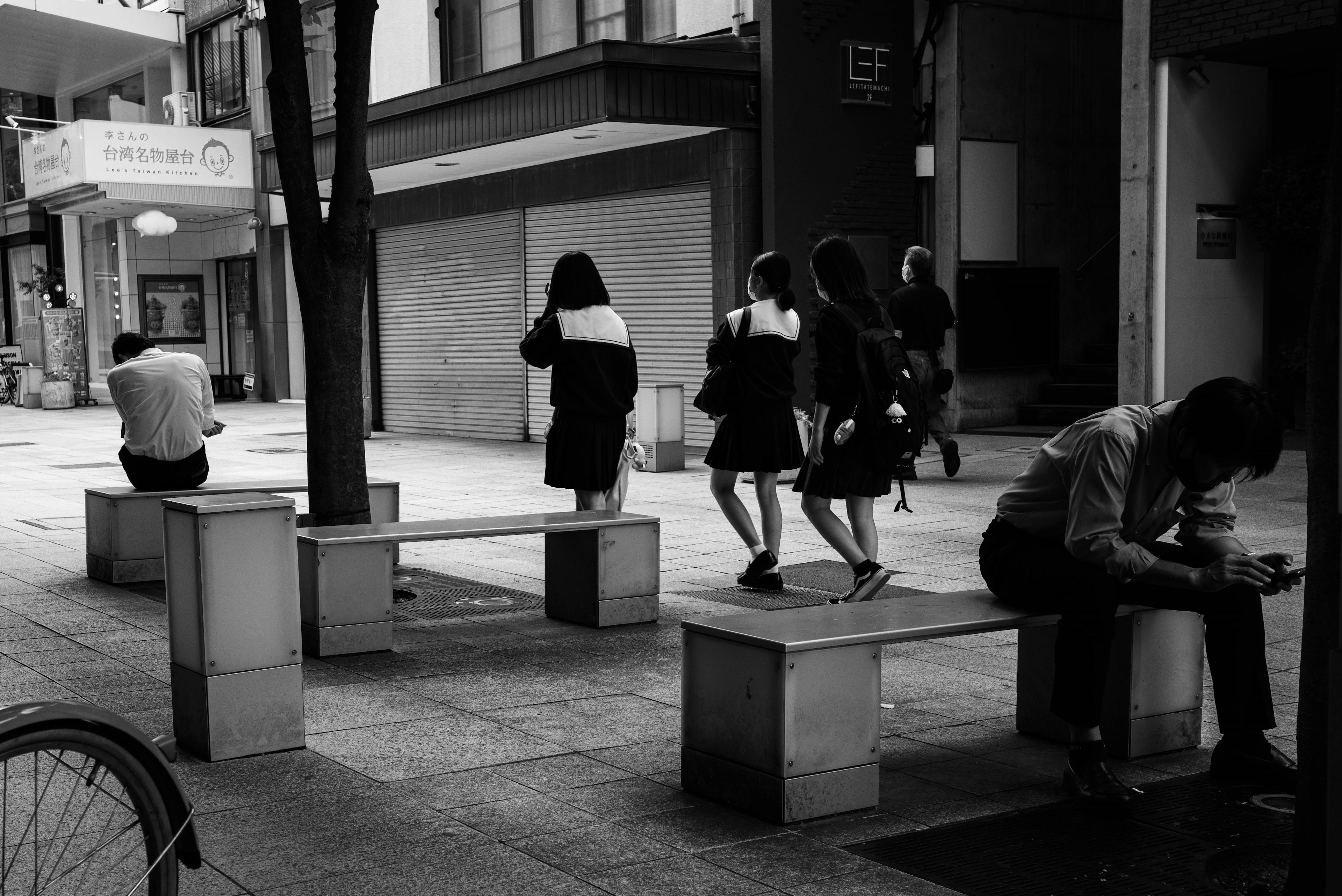 Escena urbana en blanco y negro con personas sentadas y mujeres caminando