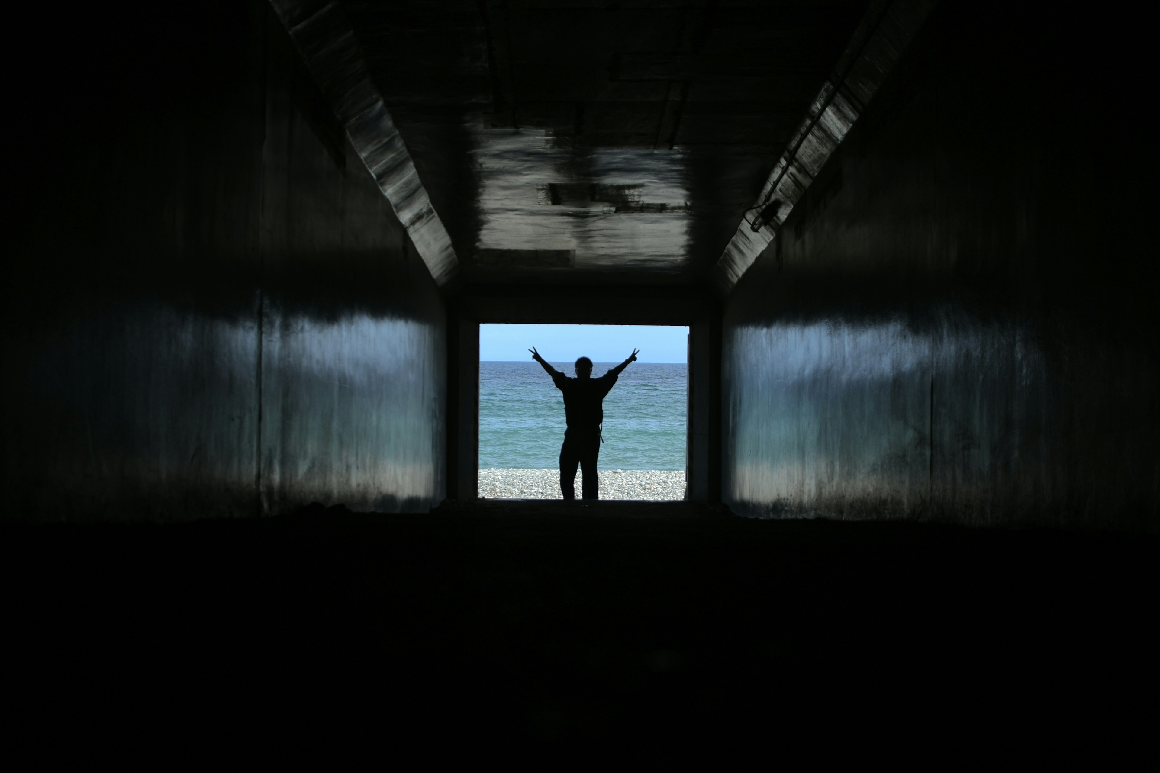 A person standing in a dark tunnel with arms wide open against a backdrop of the sea