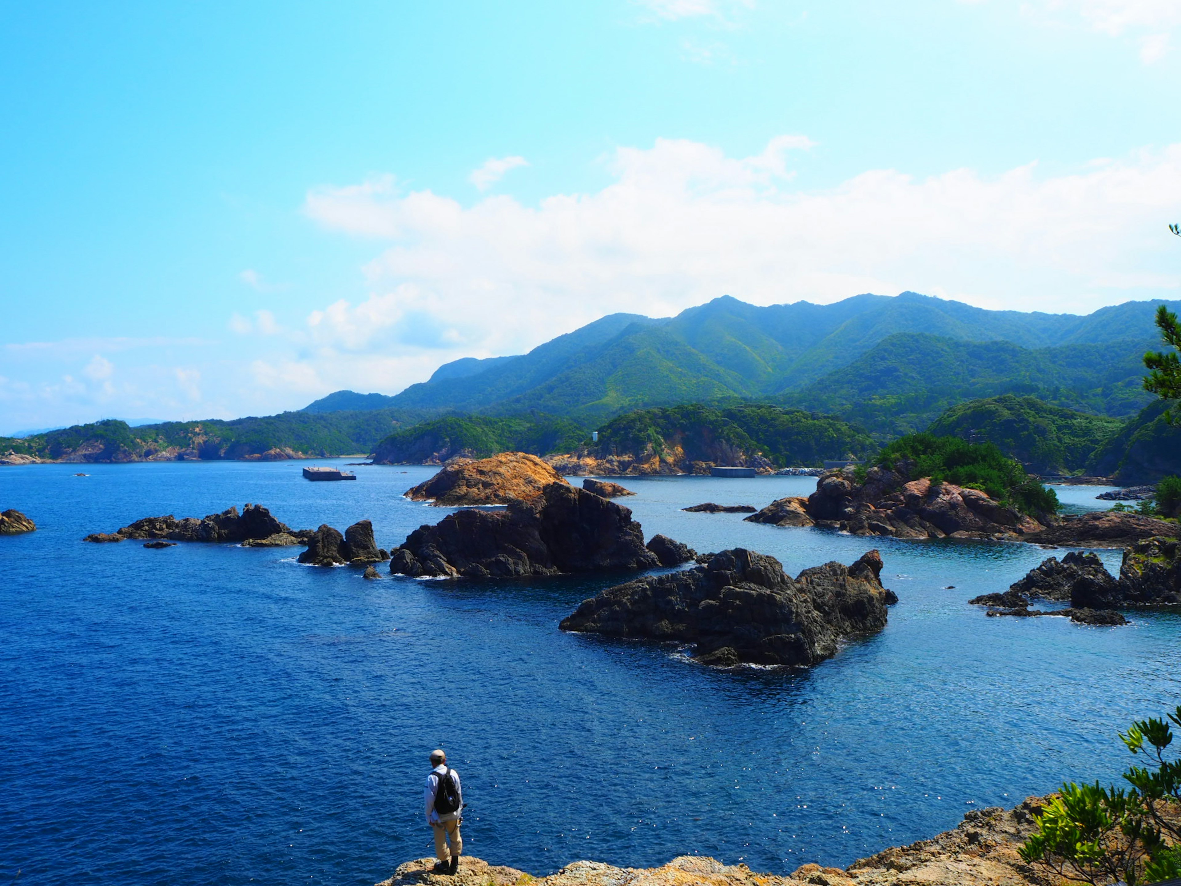 Person standing by the blue sea with green mountains in the background