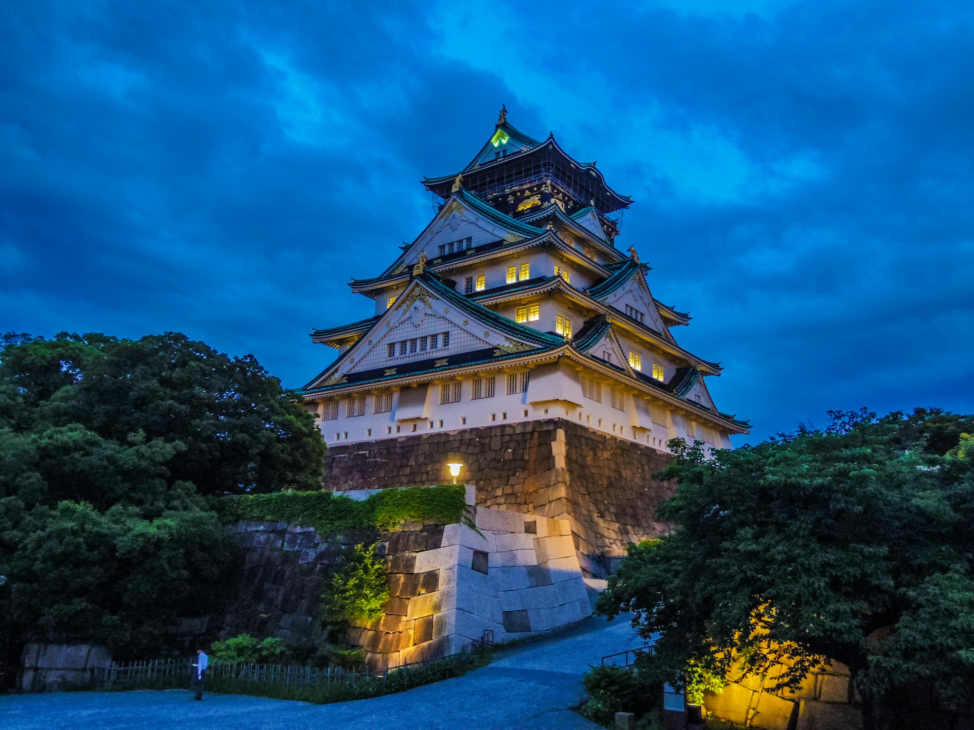 Osaka Castle illuminated at night with a dramatic blue sky and surrounding greenery