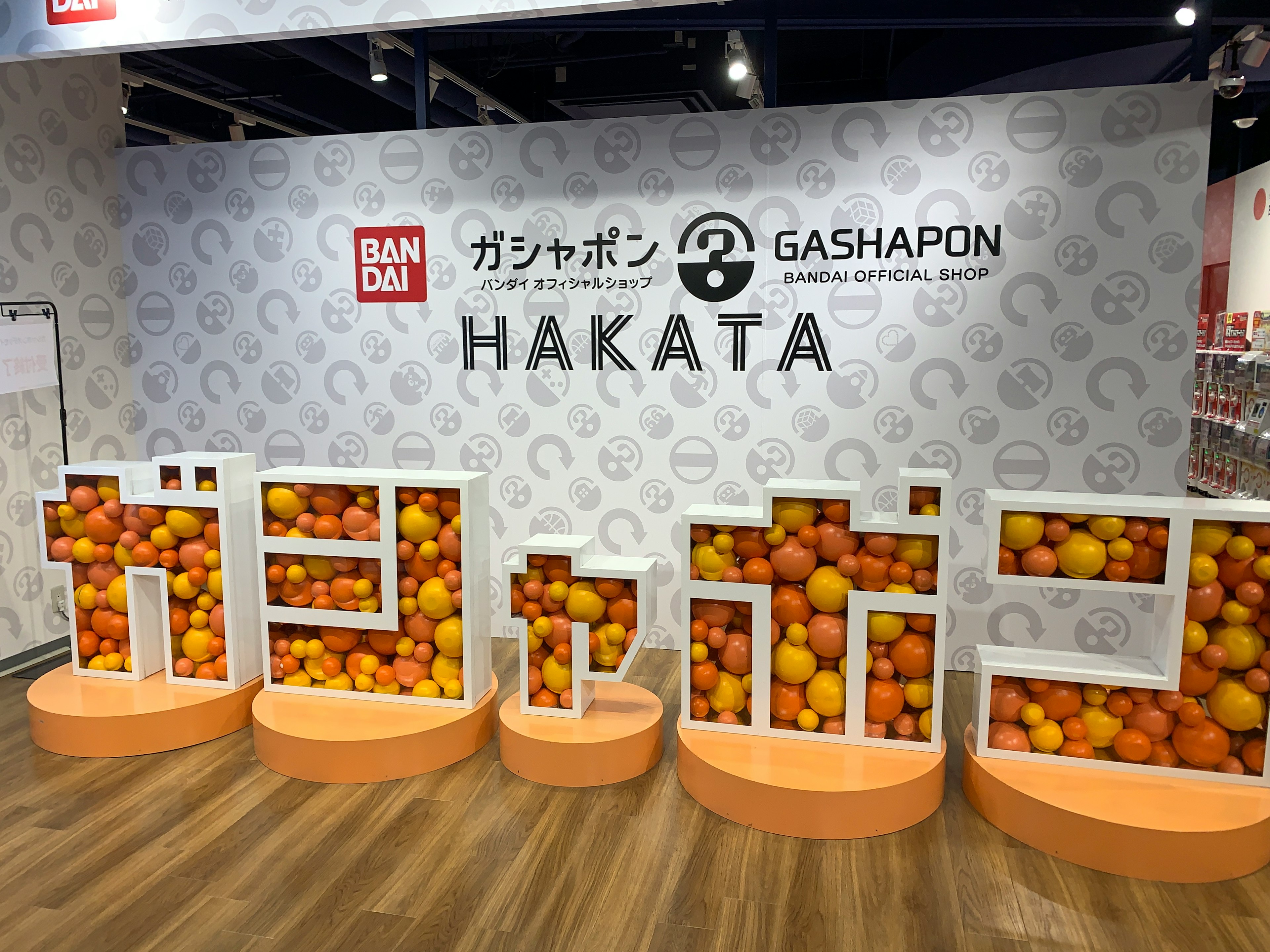 バンダイのガシャポンのディスプレイで、オレンジ色のボールが入った白い文字で「ガシャポン」と「HAKATA」の看板