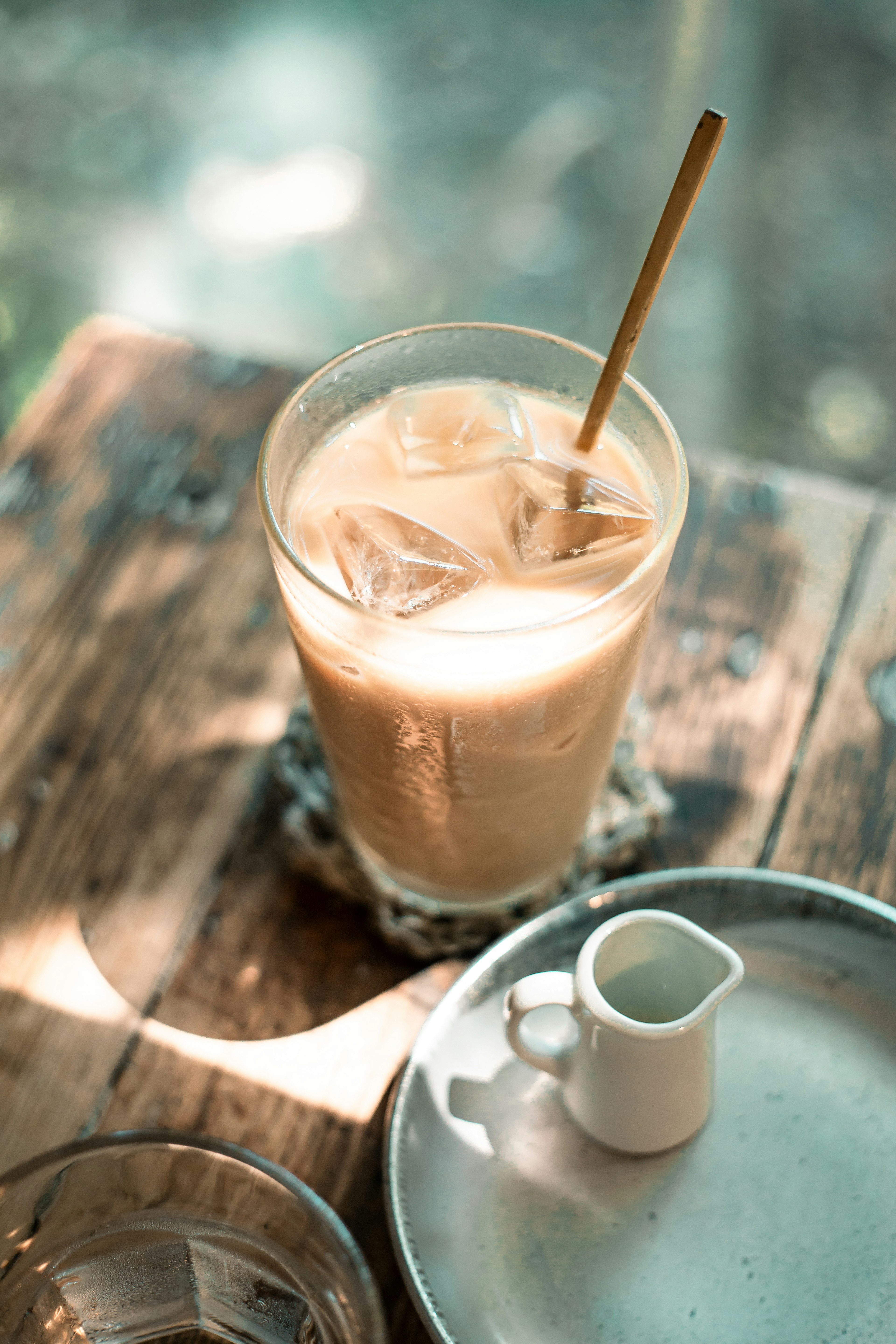 A clear glass of iced coffee with a straw and a small milk pitcher on a wooden table