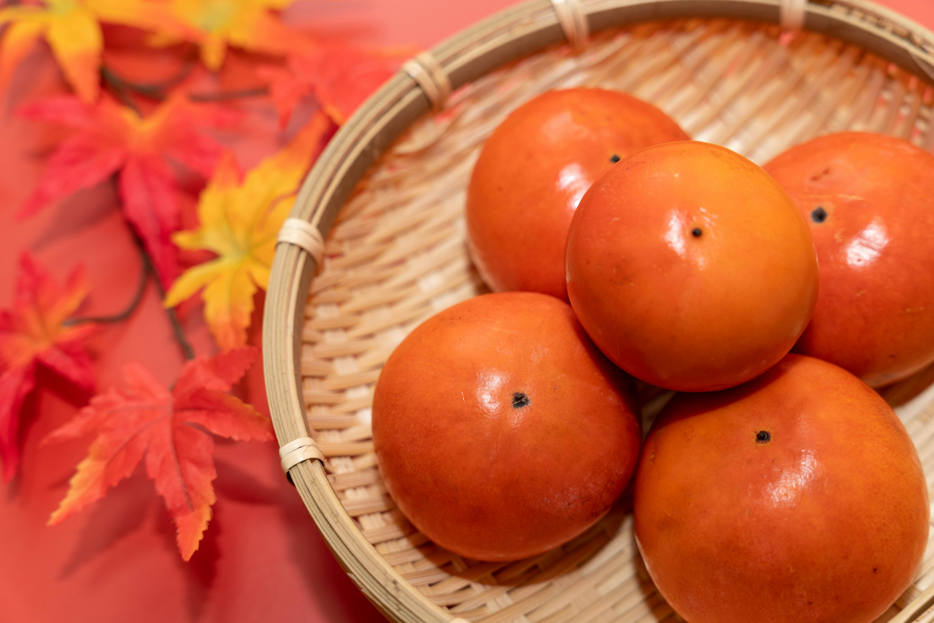 Five orange persimmons in a woven basket with autumn leaves