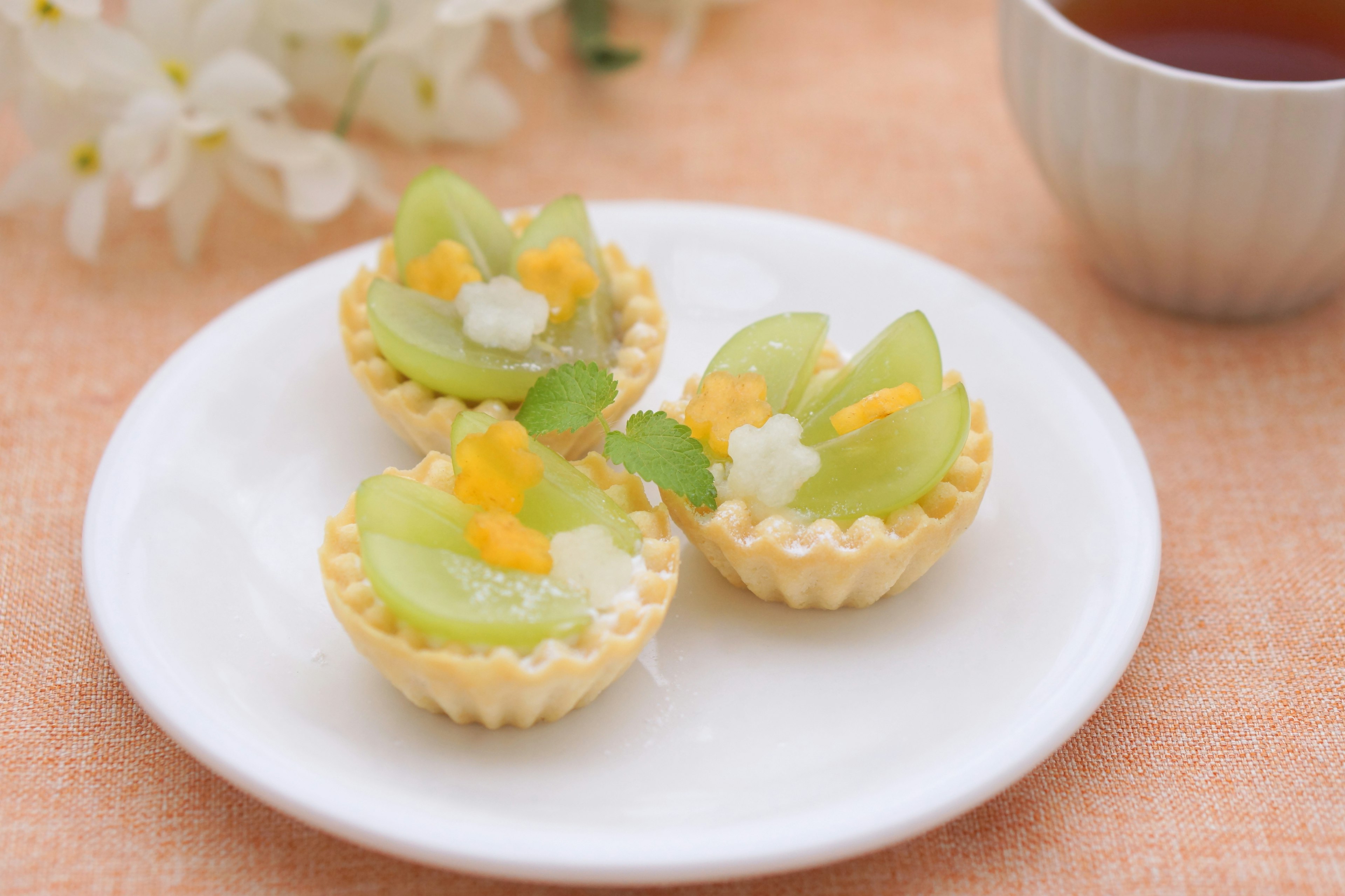 Tiga mini tart yang dihiasi dengan buah hijau dan hiasan putih di atas piring putih