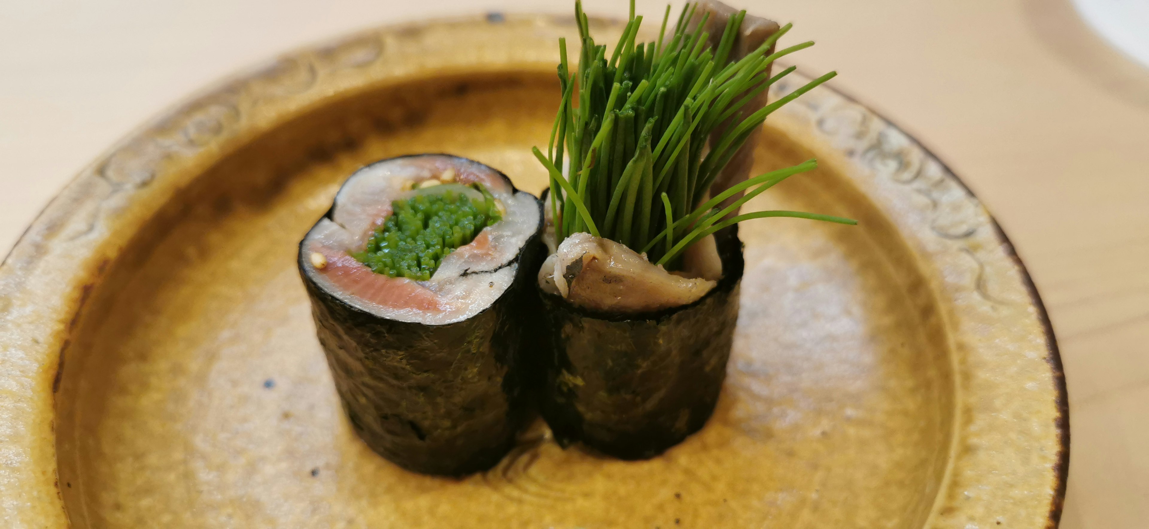 Dua gulungan sushi disajikan di atas piring dihiasi dengan daun chives hijau