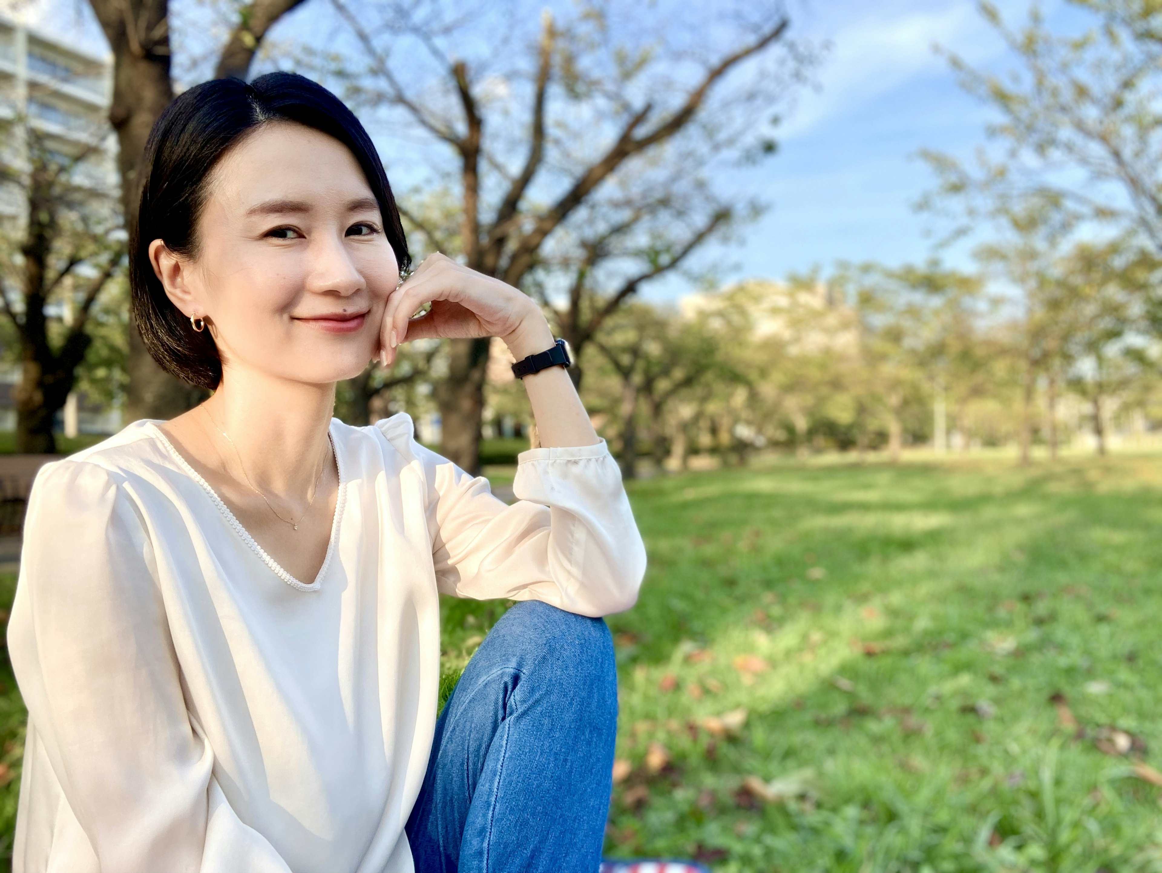 Mujer sonriente en un parque con blusa blanca y jeans azules césped verde y árboles de fondo