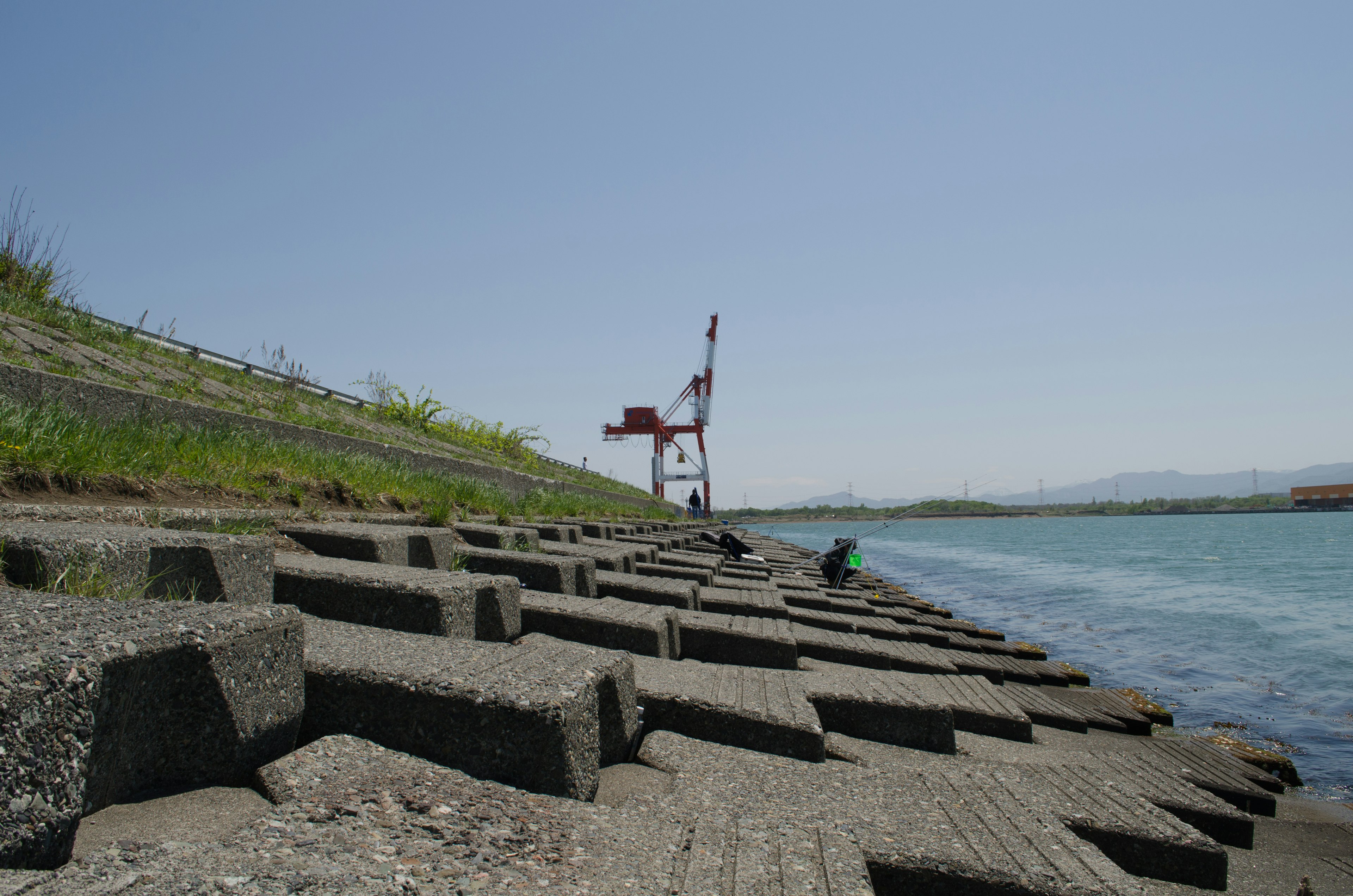 コンクリートブロックが並ぶ岸辺と遠くにクレーンが見える河川の風景