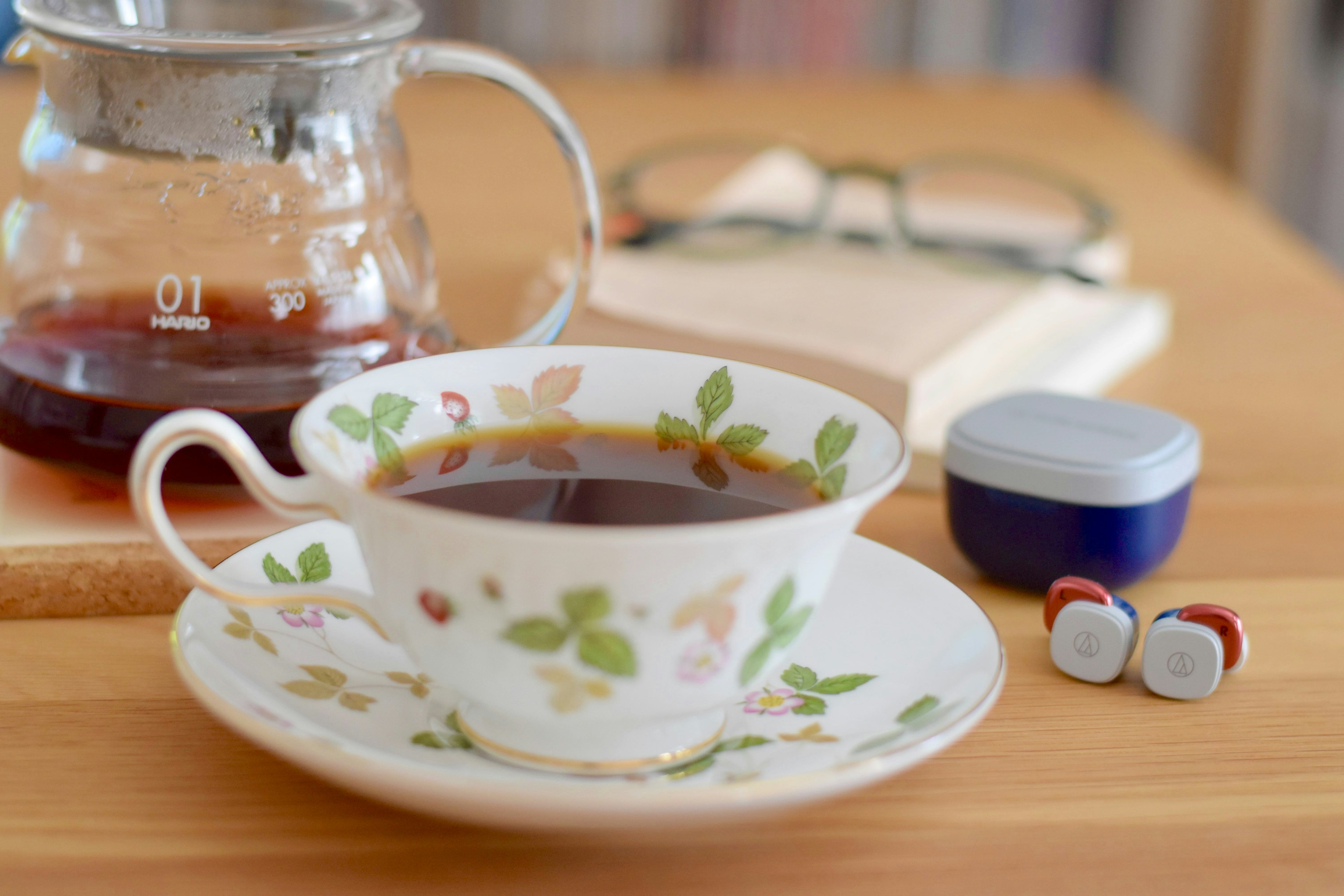 Una tazza da tè floreale riempita di caffè accanto a una caffettiera con auricolari e un libro