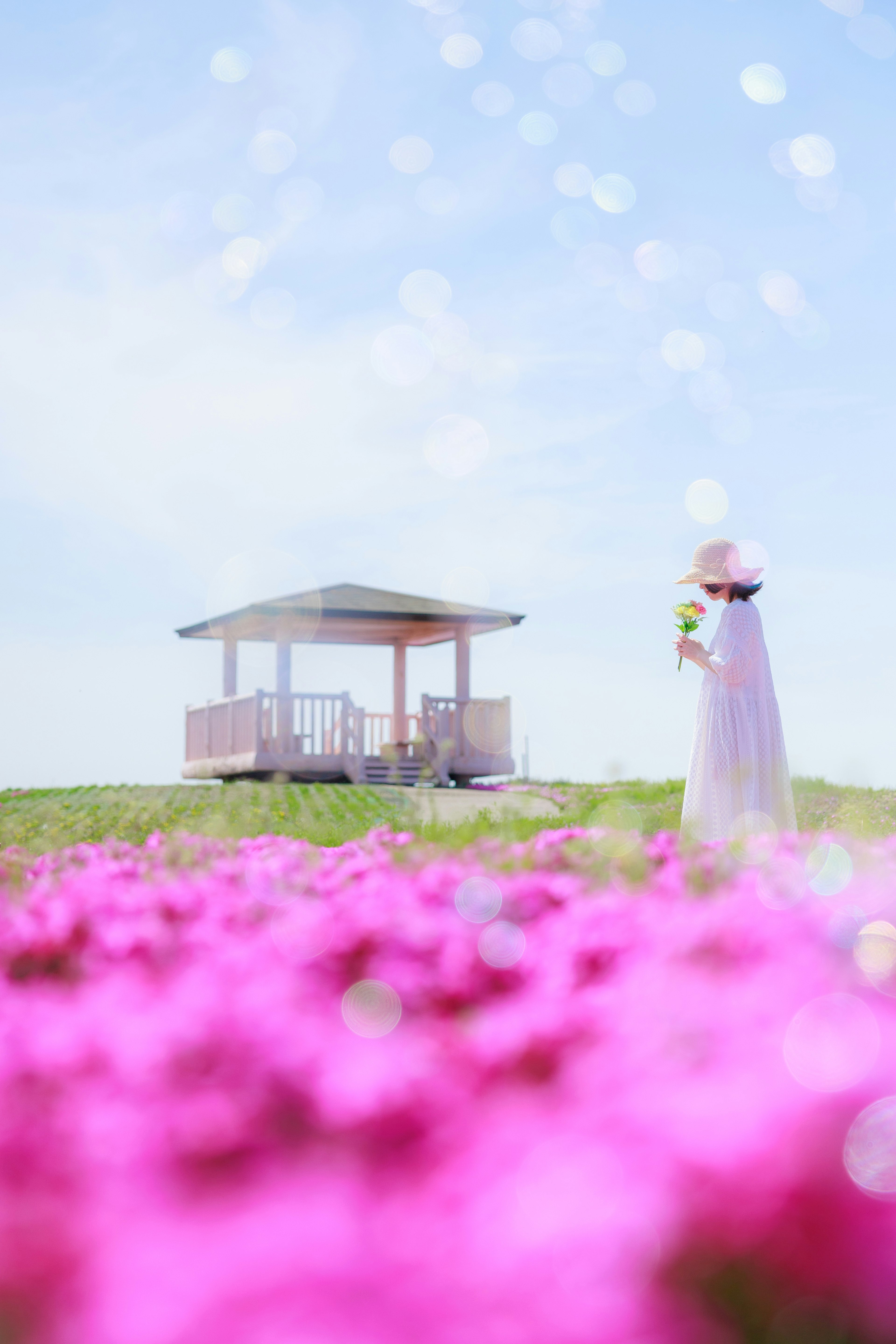 Eine Frau in einem weißen Kleid steht in einem Feld mit rosa Blumen neben einem Pavillon