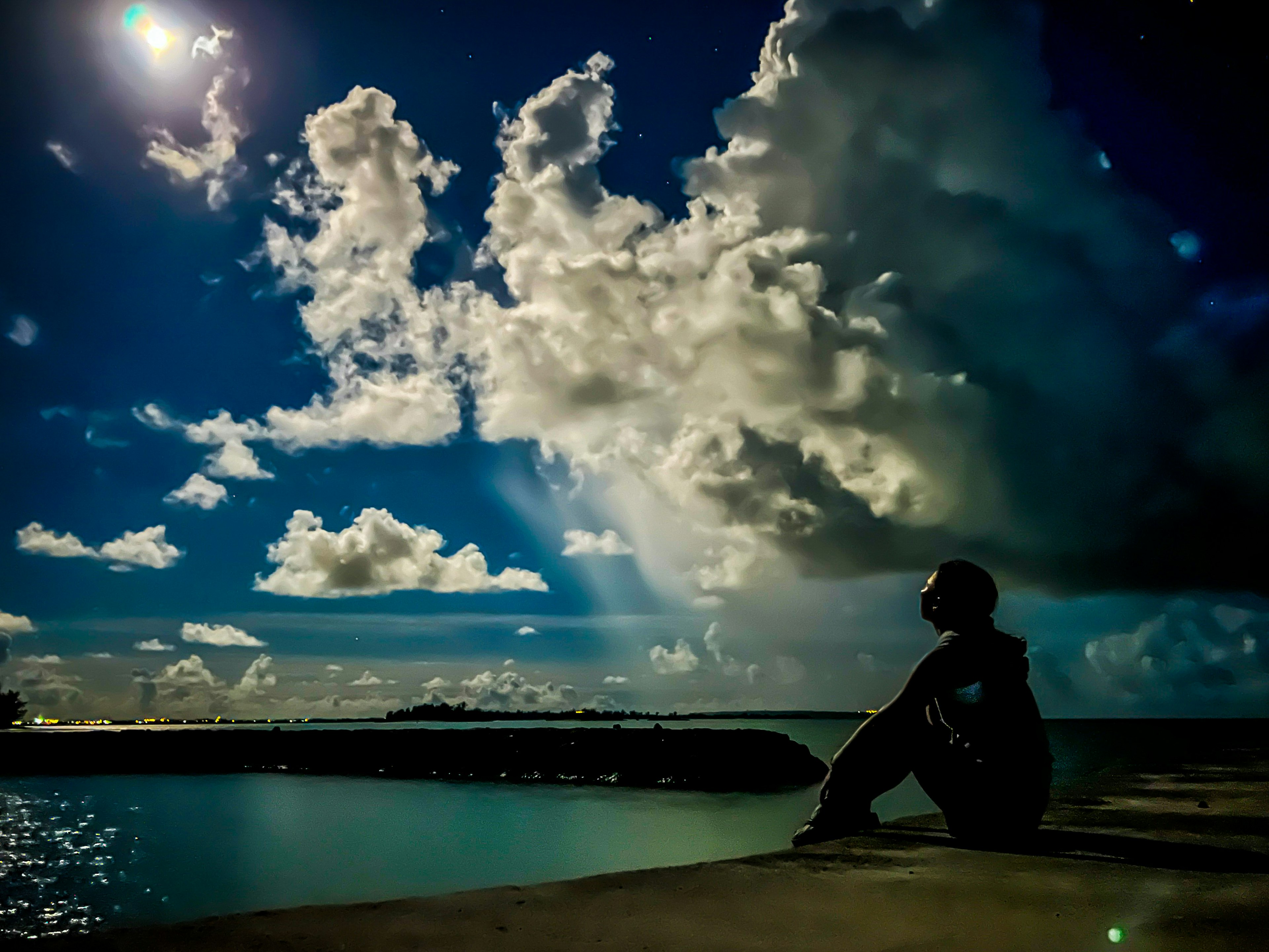 Eine Person sitzt am Wasser und schaut auf einen sternenklaren Himmel mit dramatischen Wolken