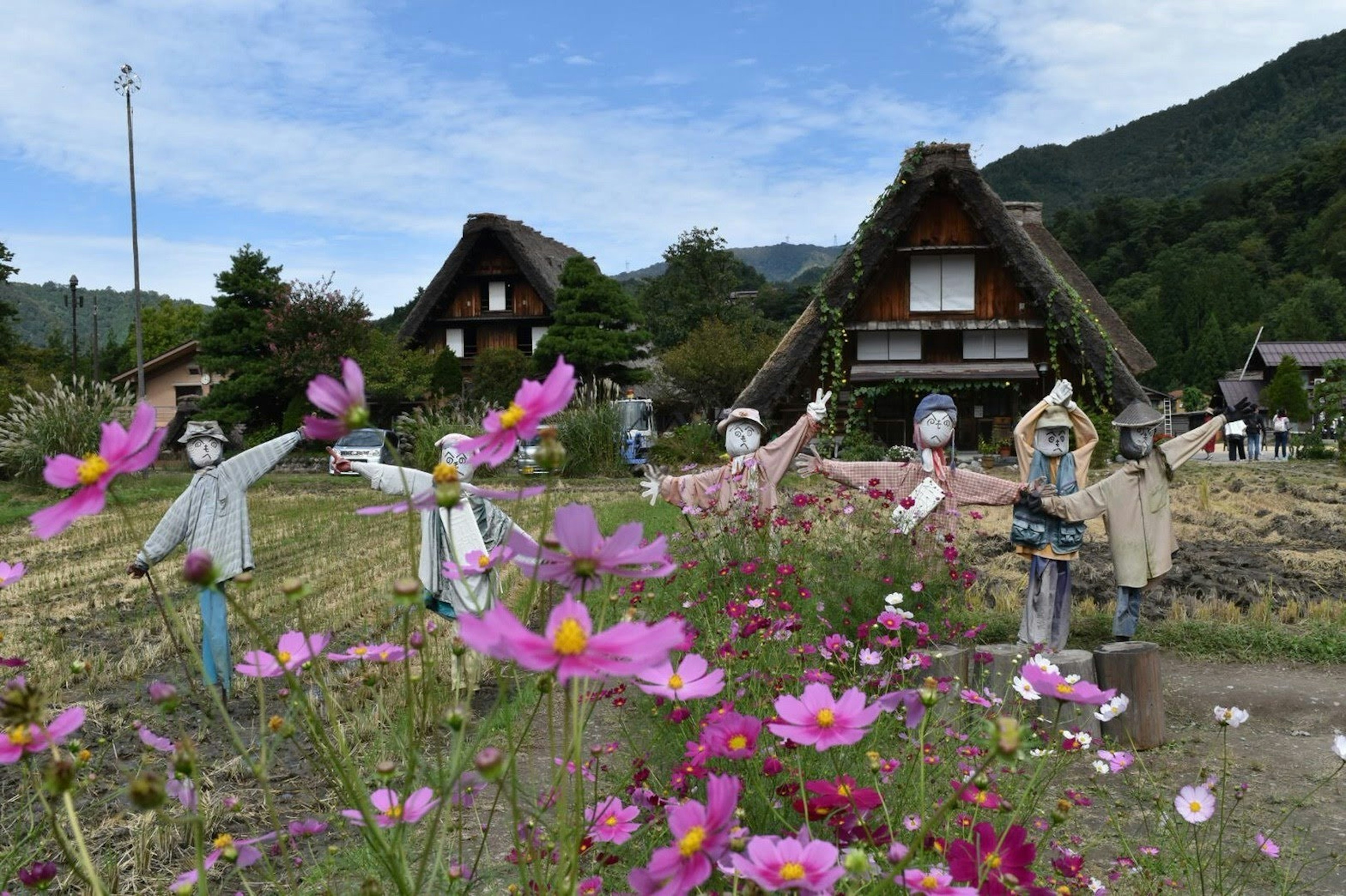 美しい花と伝統的な合掌造りの家が並ぶ風景で農作業をする人形が見える