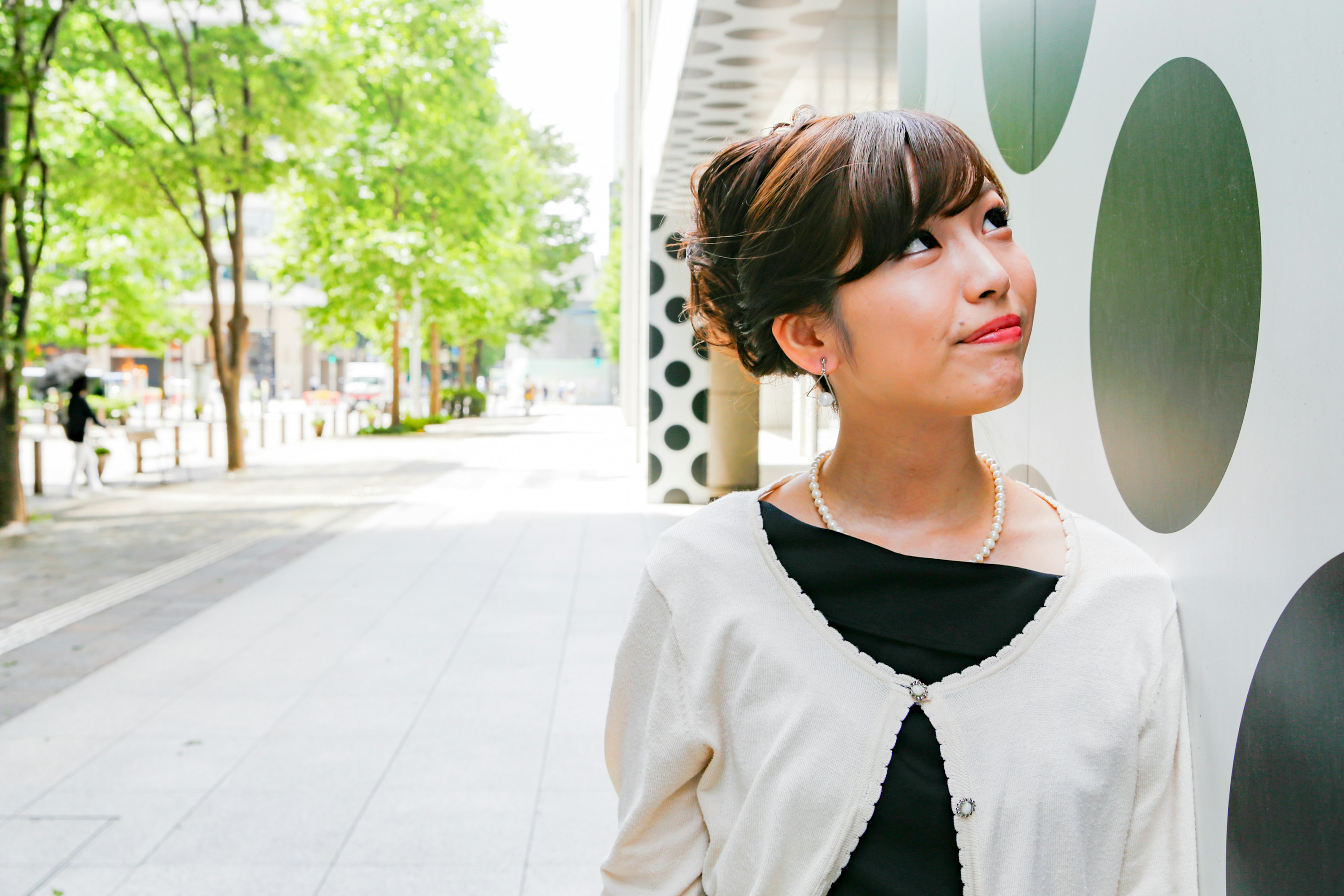 A woman posing between green trees with a modern building backdrop