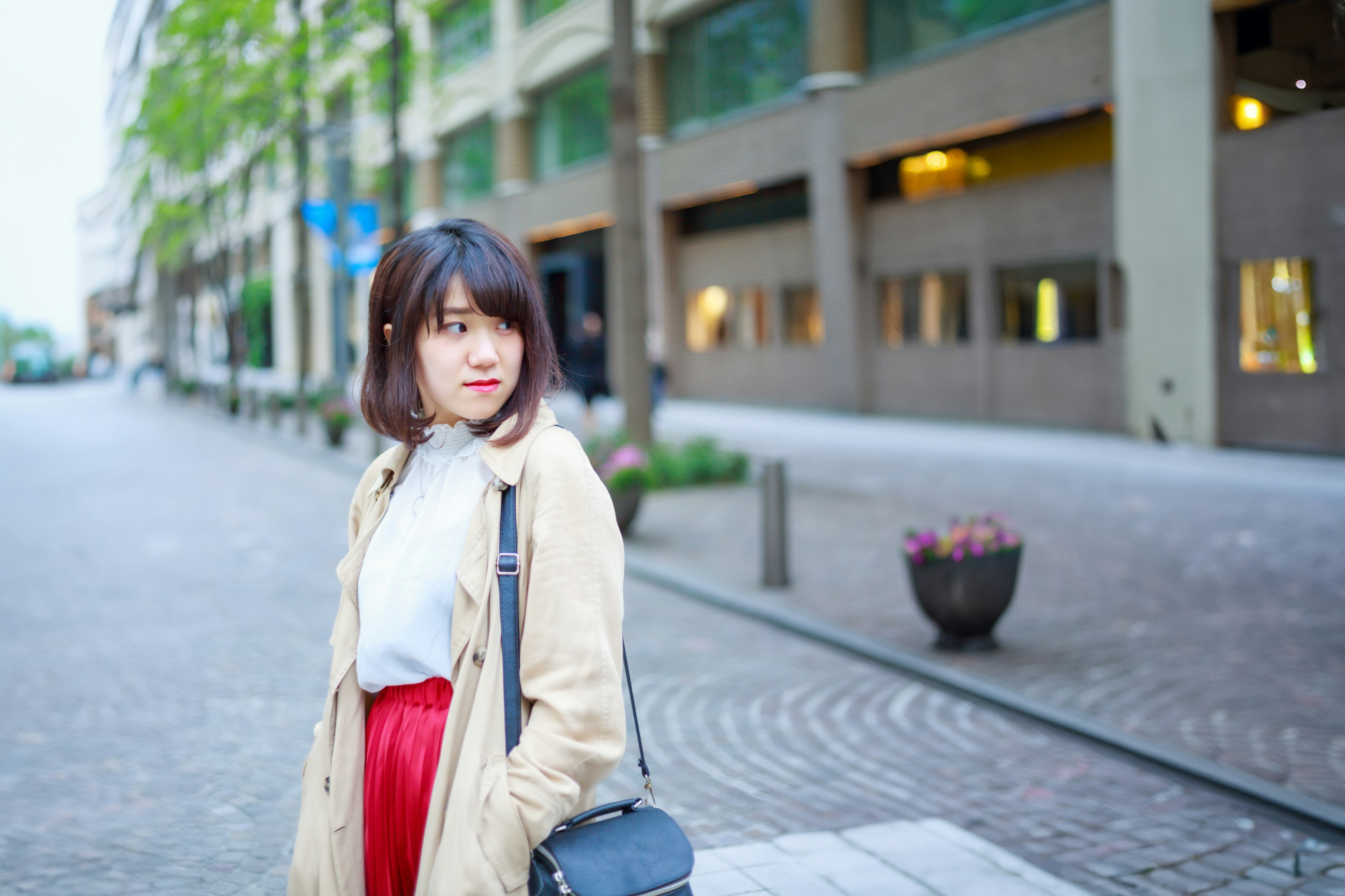 Une femme sourit dans la rue portant un manteau beige et une jupe rouge avec les cheveux courts