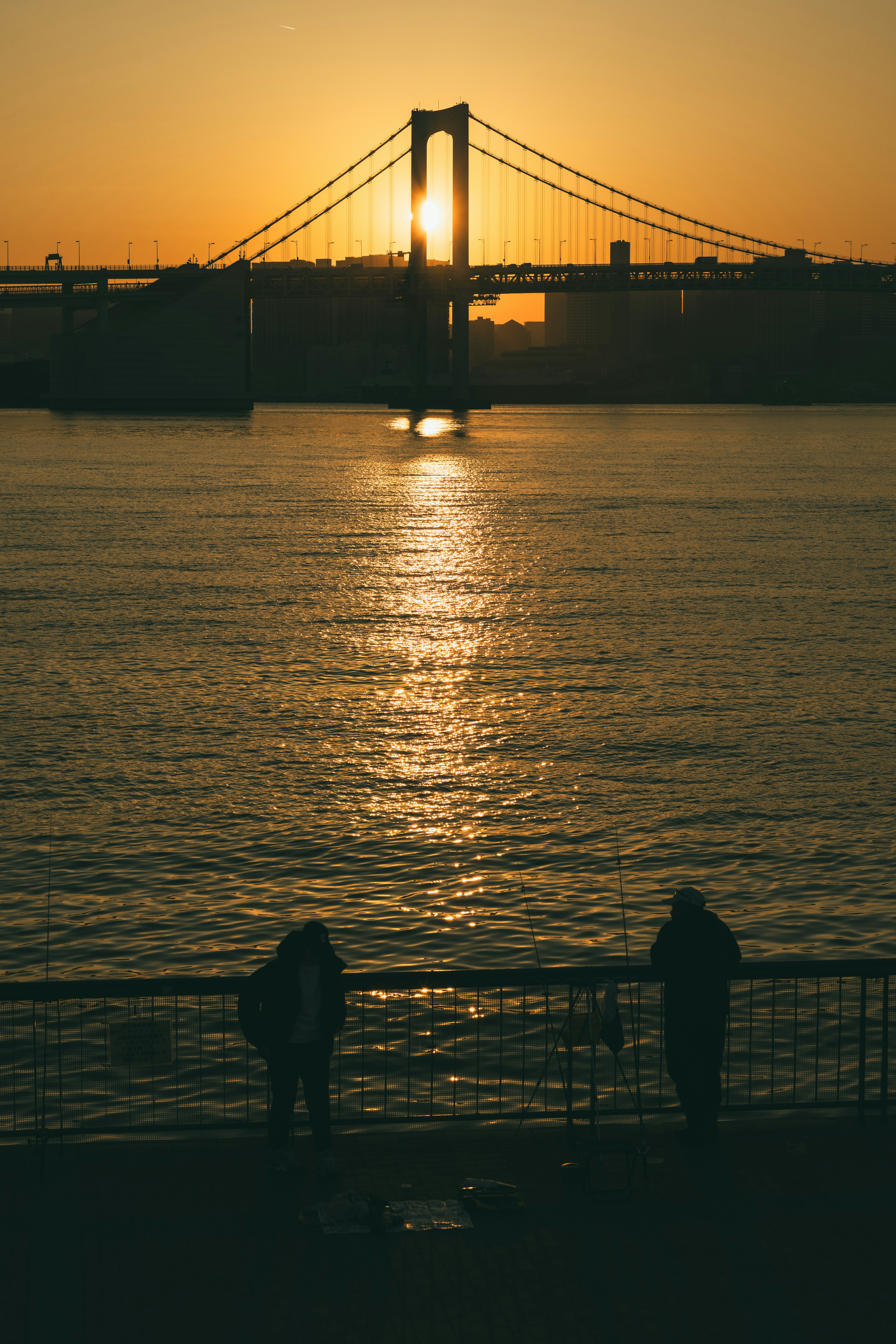 Two figures by the water at sunset with a bridge in the background