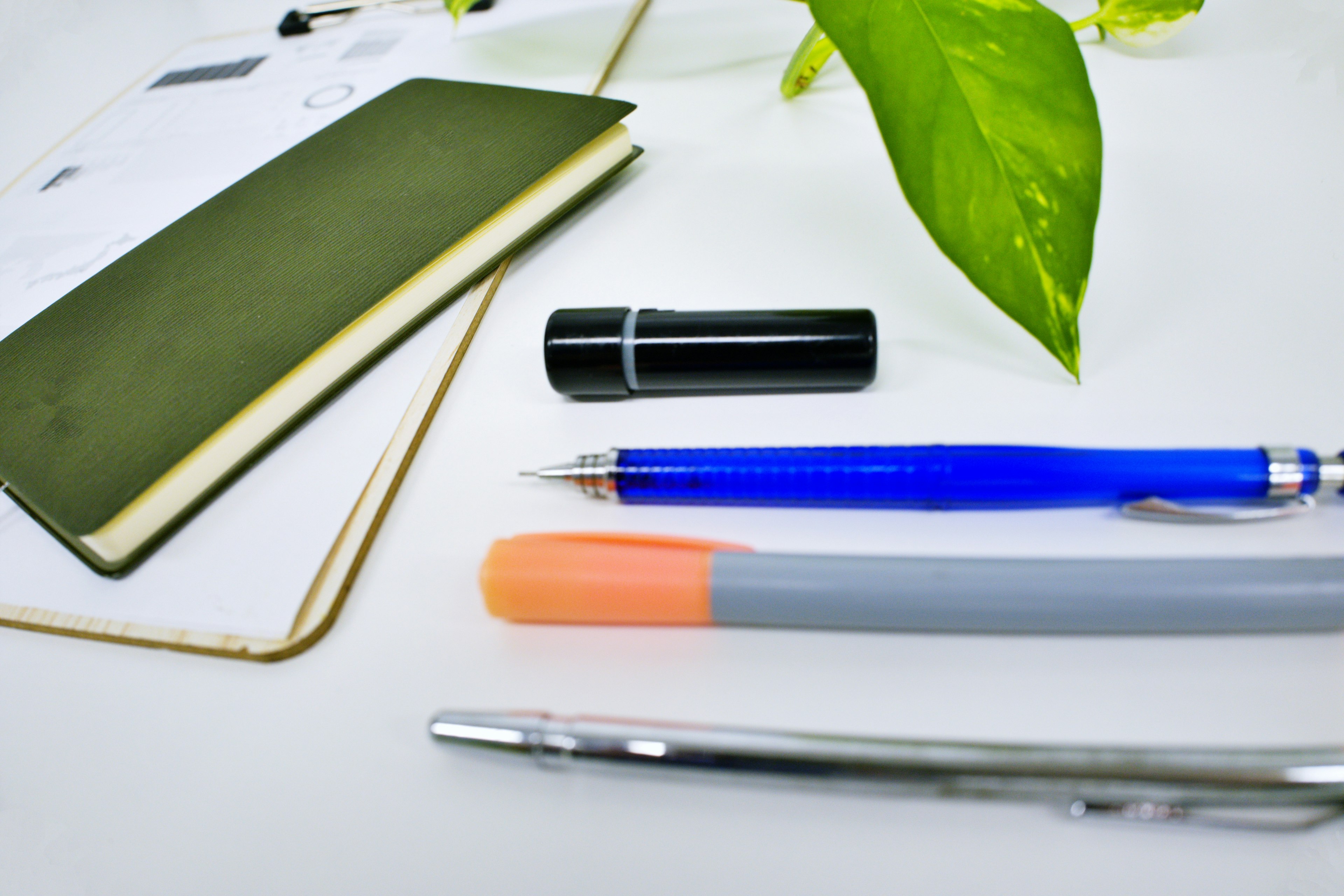 A green notebook with a black pen on a desk alongside a blue ballpoint pen and an orange marker