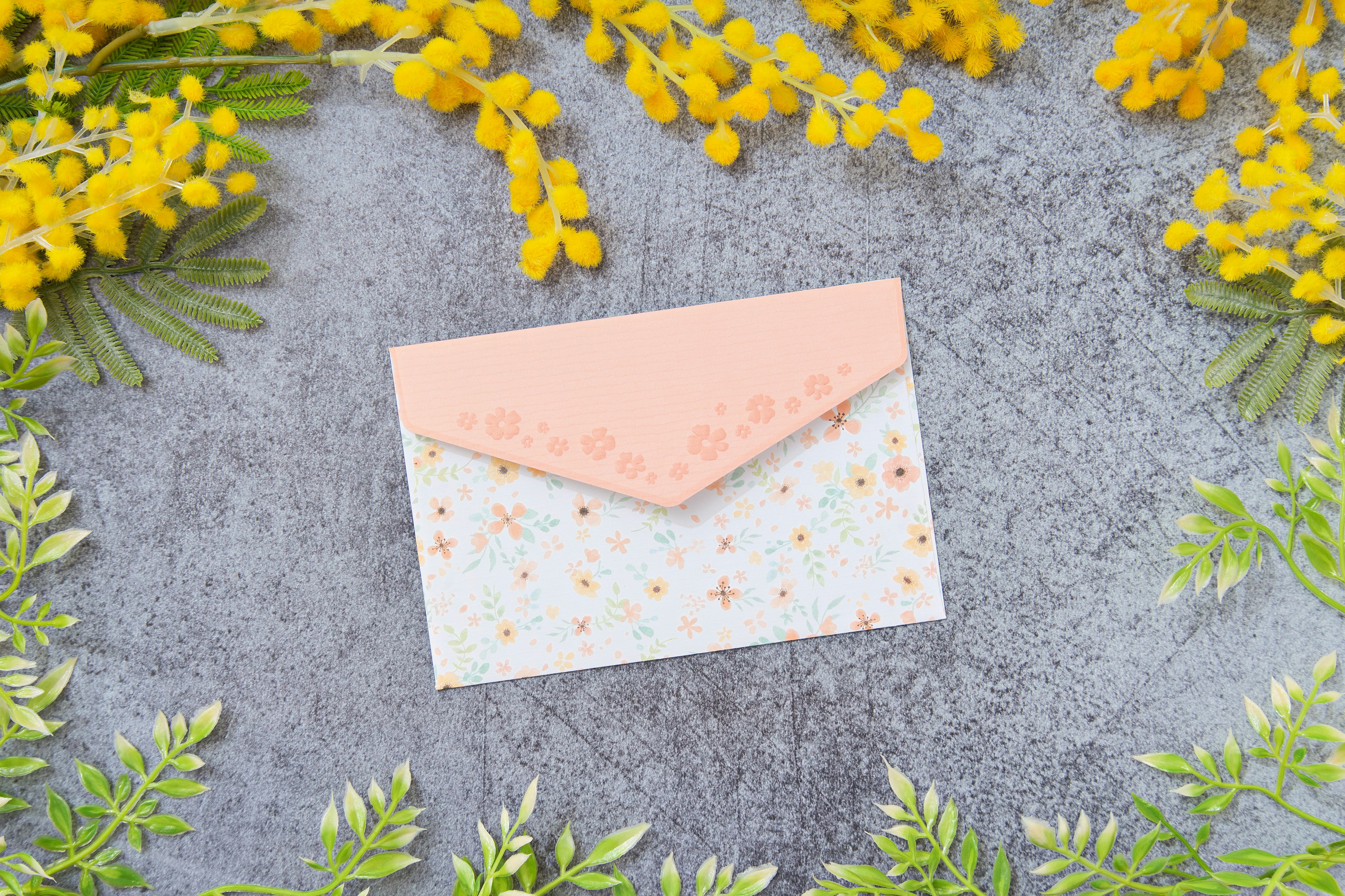 A pink envelope on a gray background surrounded by yellow flowers and green leaves